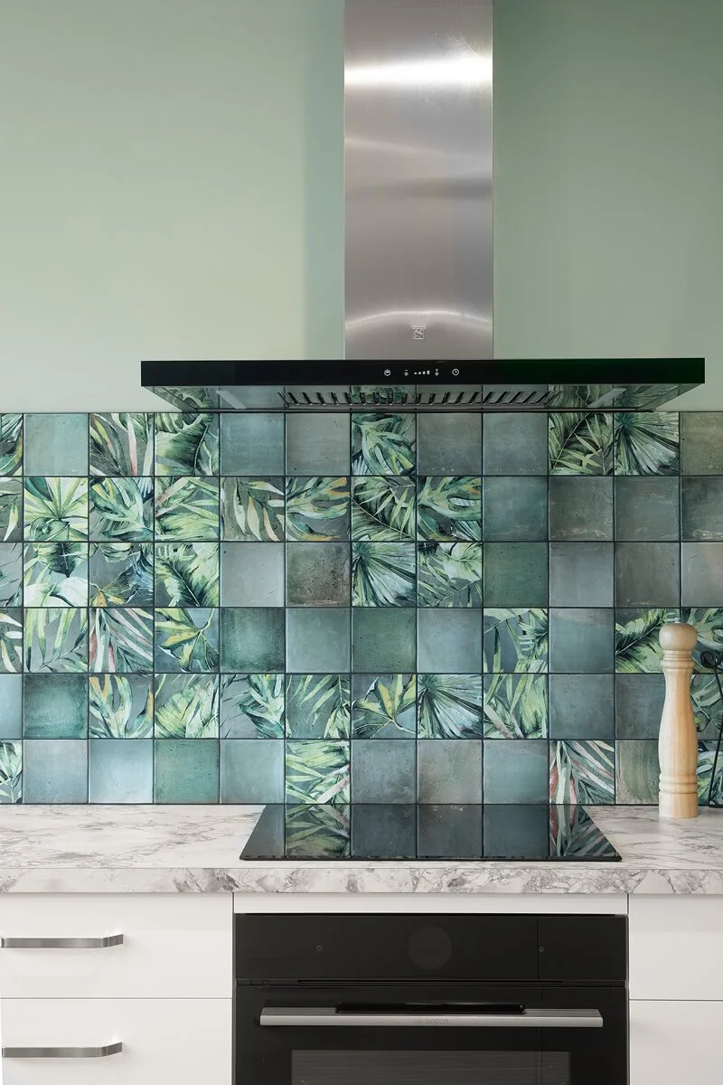 Green tiled kitchen splashback with botanical patterned tiles, marble-look benchtop and black cooktop.