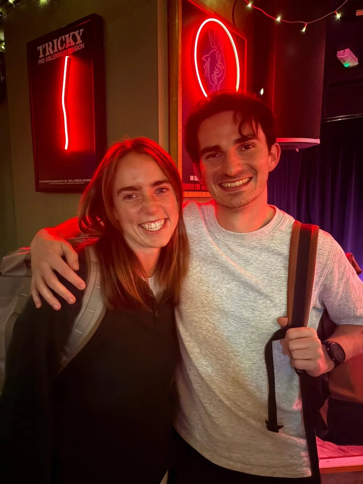 A smiling woman with red hair and a smiling man with dark hair, both in casual clothing, standing close together in a dimly lit venue with neon red signs and string lights.
