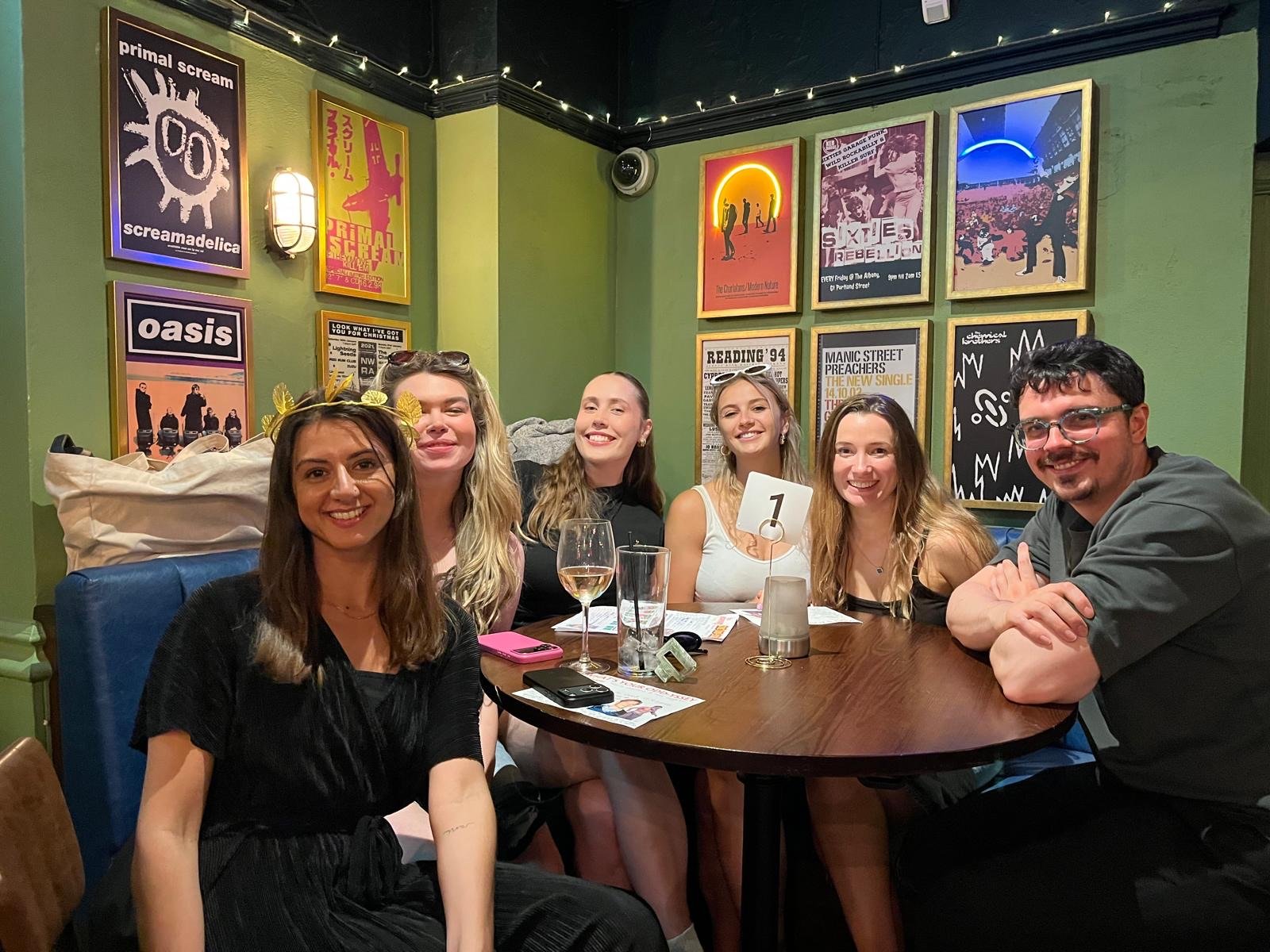 Six friends sitting at a round table in a restaurant, smiling and posing for a photo. There are drinks including wine and water, and various menus and items on the table. The background features green walls adorned with colorful posters of different 