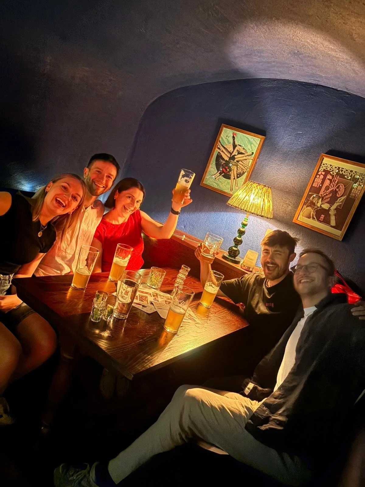 Group of five friends sitting at a wooden table in a cozy, dimly lit pub or bar, raising glasses in a toast and smiling, with various empty beer glasses and shot glasses on the table. The walls are painted dark blue and decorated with colorful framed artwork and a small table lamp.