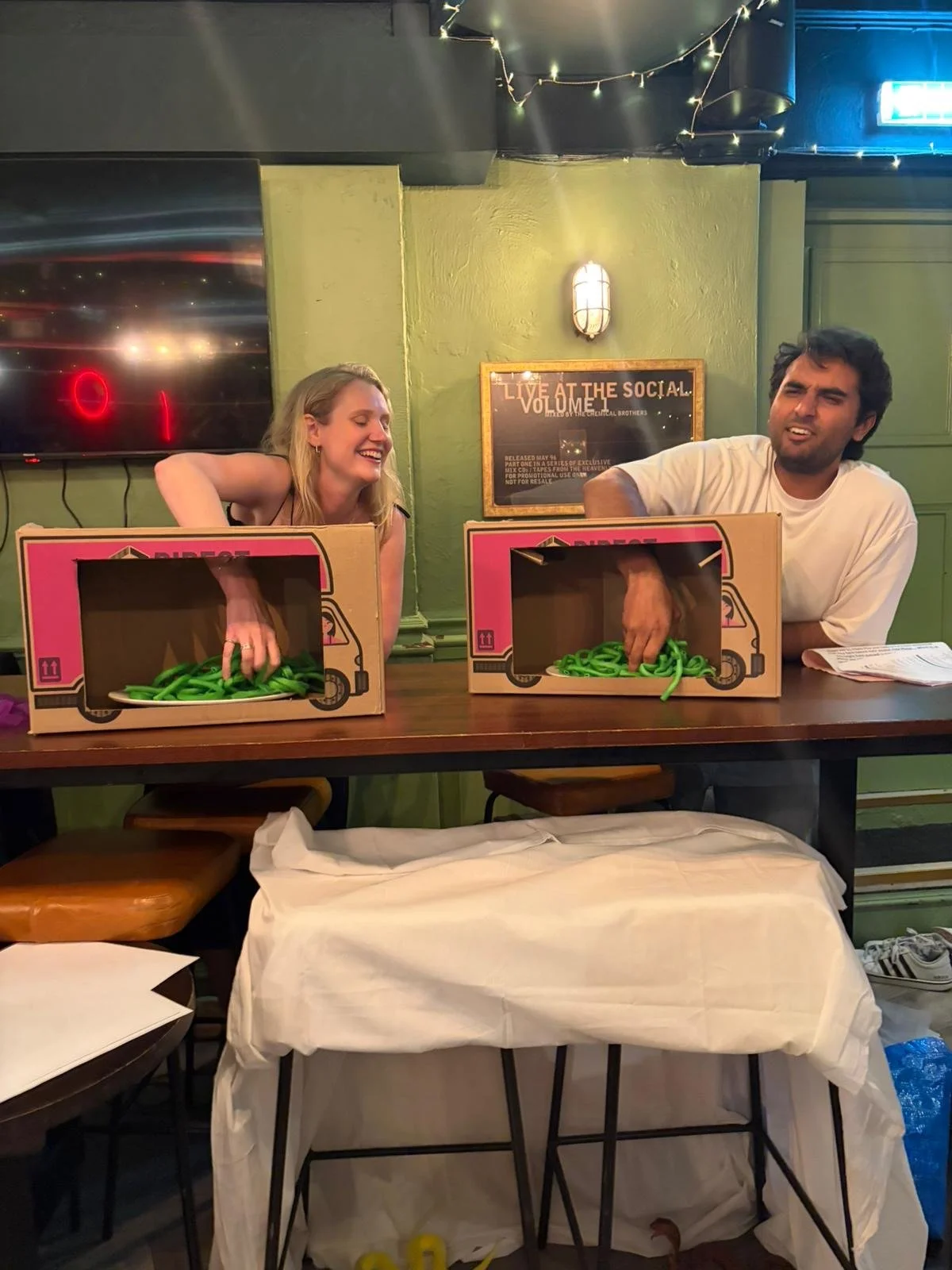 Two people, a woman and a man, are sitting at a table with cardboard boxes filled with green rubber band-like objects. The woman is smiling and the man is making a funny face. The background includes a TV, a wall sign, and string lights.