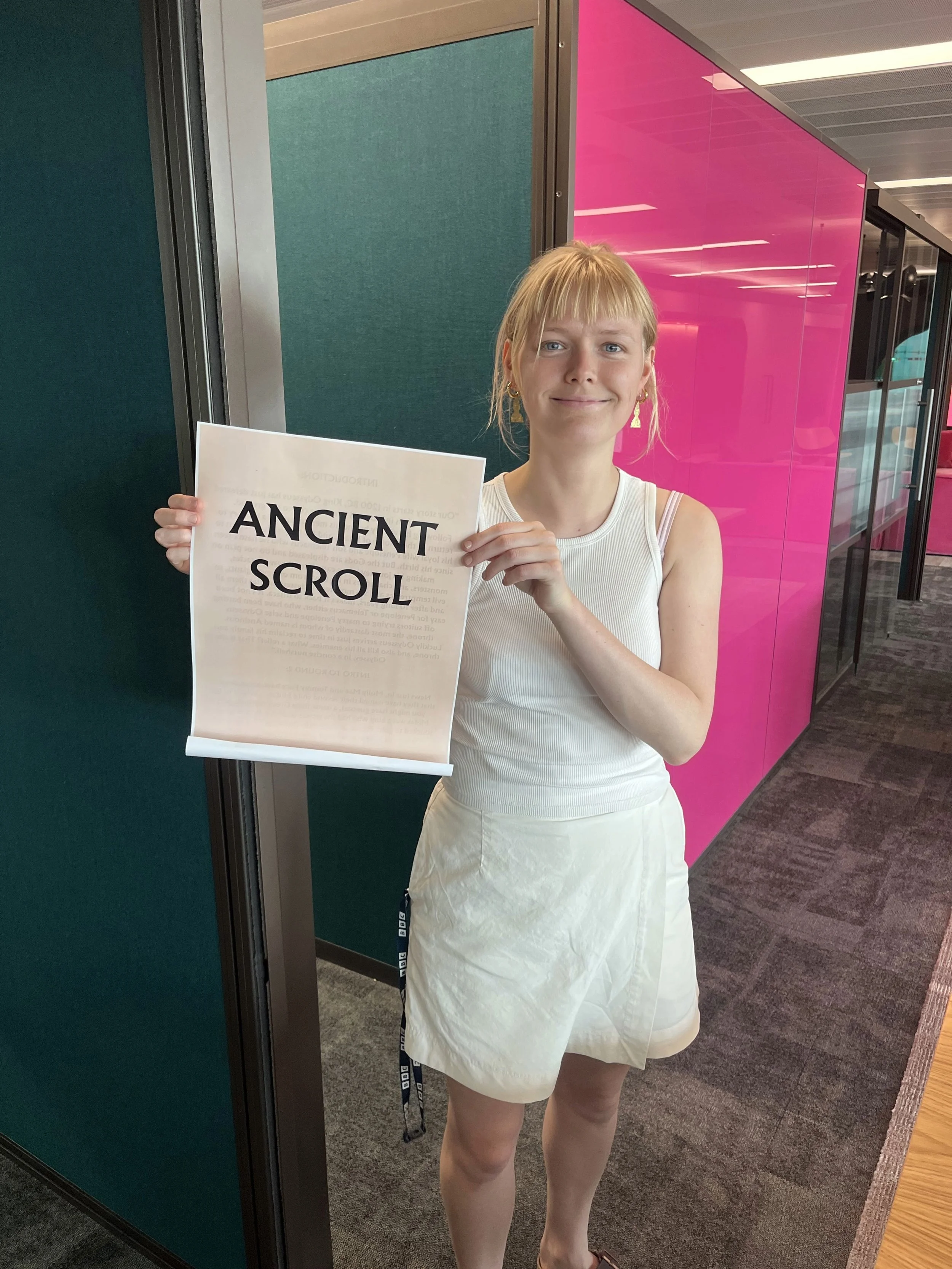 A woman standing in an indoor space holding a sign that reads 'Ancient Scroll'. She is wearing a white sleeveless top and a white skirt with a slit, and has blonde hair. The background includes green and pink walls and gray carpeting.