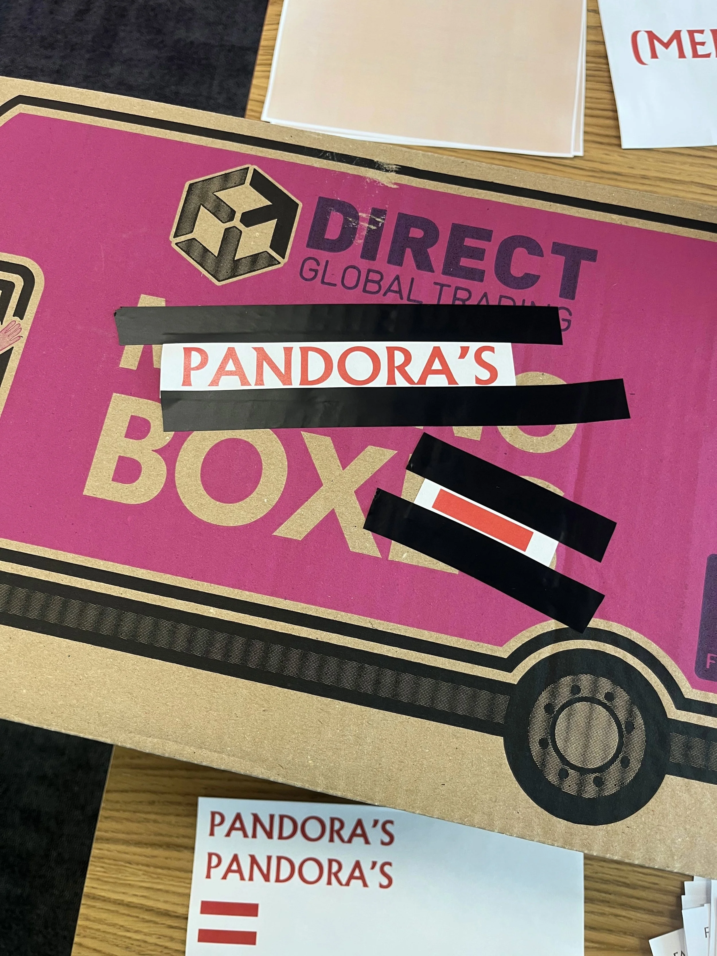 A pink cardboard box with the words "DIRECT GLOBAL TRADING" and a logo above. Blacked-out labels partially cover a white label with red text that says "PANDORA'S." The box is on a wooden surface with some papers and other objects nearby.