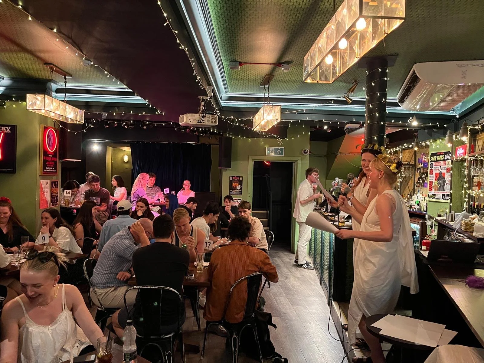 People dining in a lively restaurant or bar with festive lighting. Two women in white dresses are speaking or singing at the bar, and others are seated at tables, engaged in conversations.