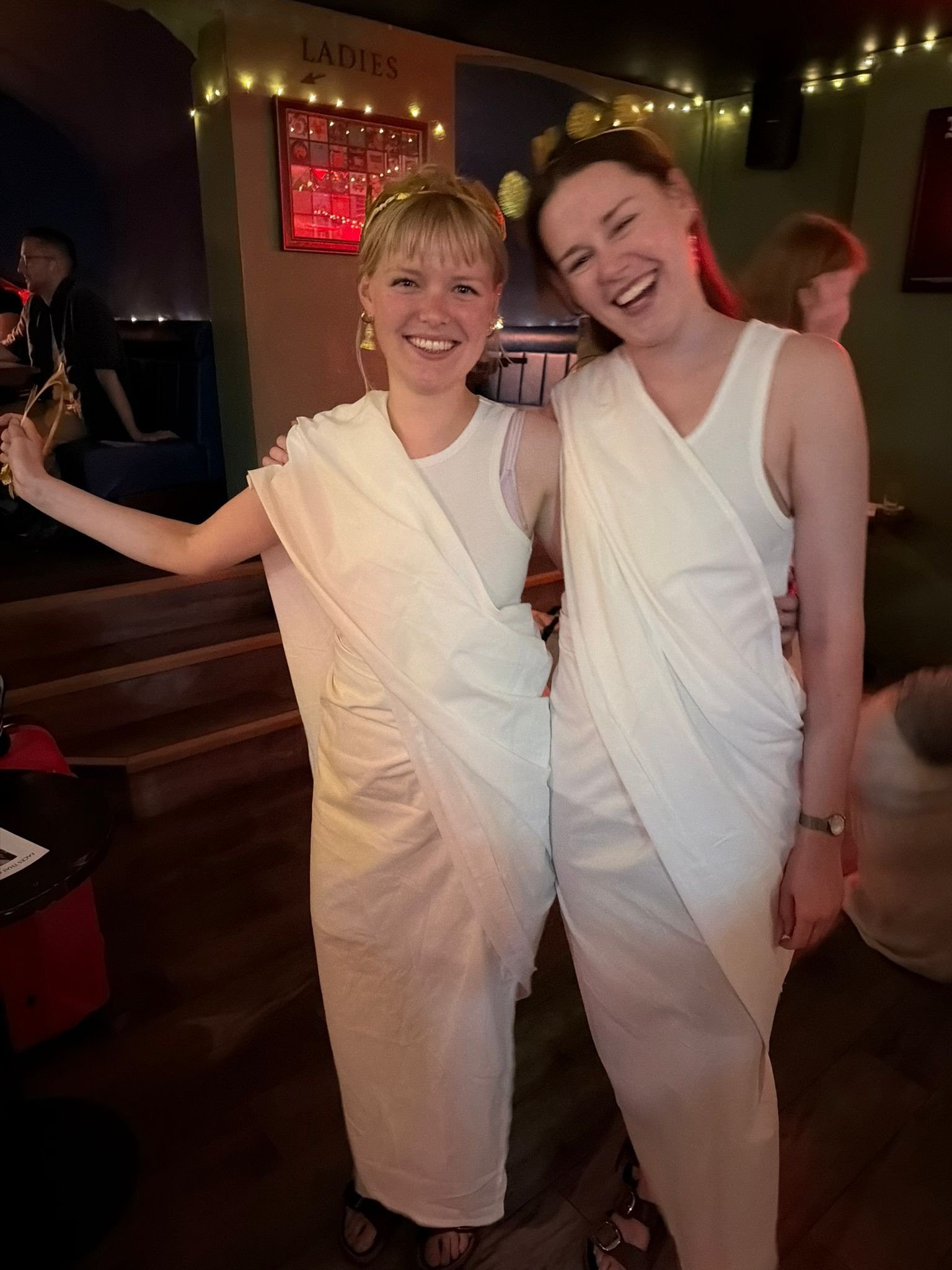 Two young women dressed in white toga costumes at a party or celebration, smiling and posing for the photo, with a sign indicating 'Ladies' restroom in the background.