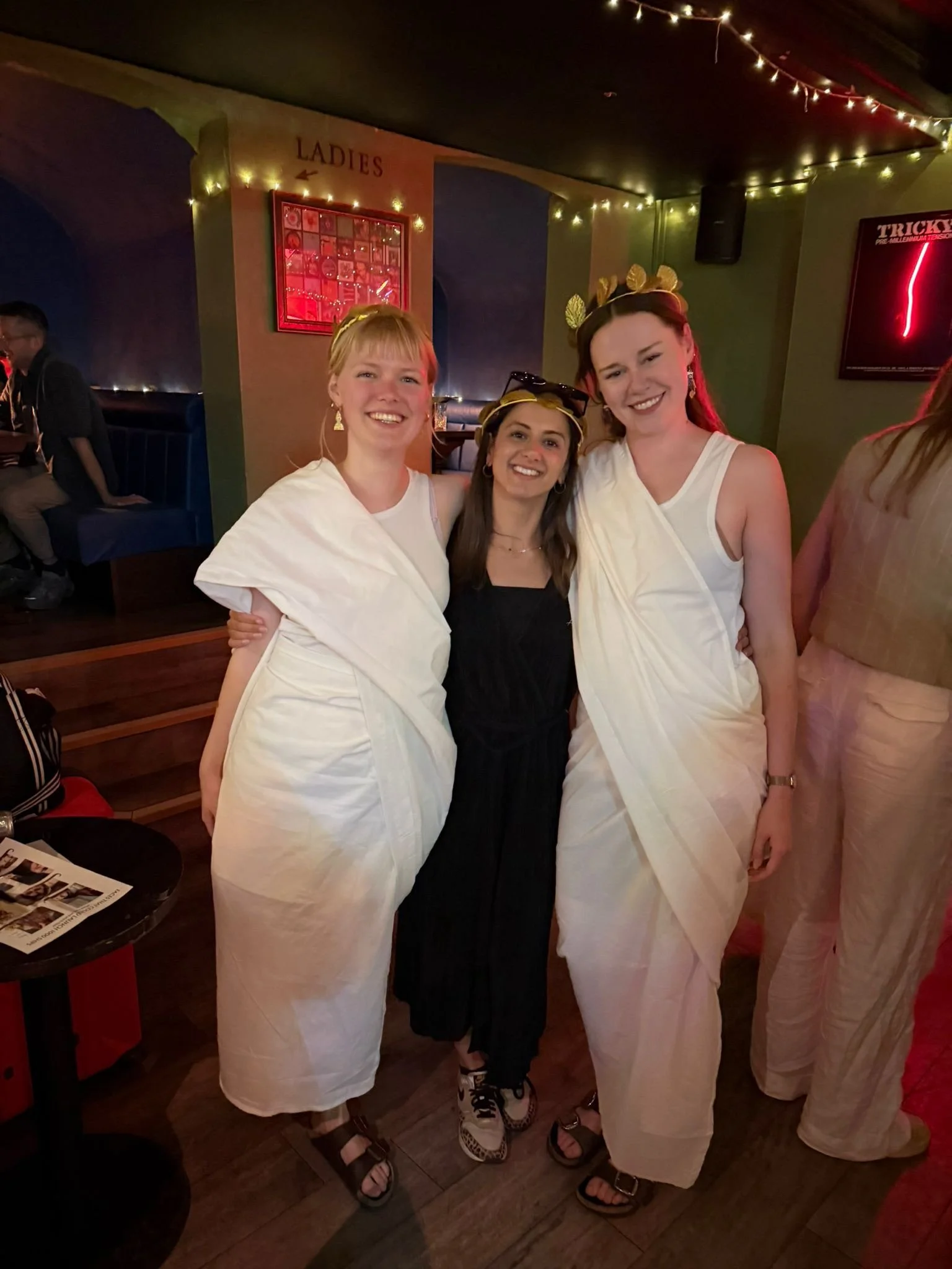 Three women smiling and wearing white dresses and gold headbands with leaf motifs, standing together in a dimly lit room with string lights. Two are embracing slightly, and one is wearing sunglasses on her head.