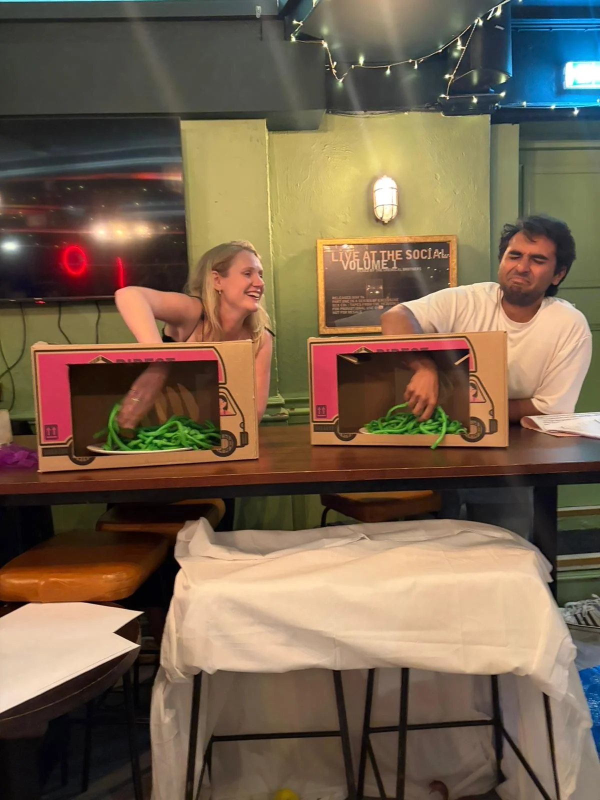 Two people sitting behind a table with pink boxes of realistic-looking fake green noodles, pretending to play with them as if they are actual food.