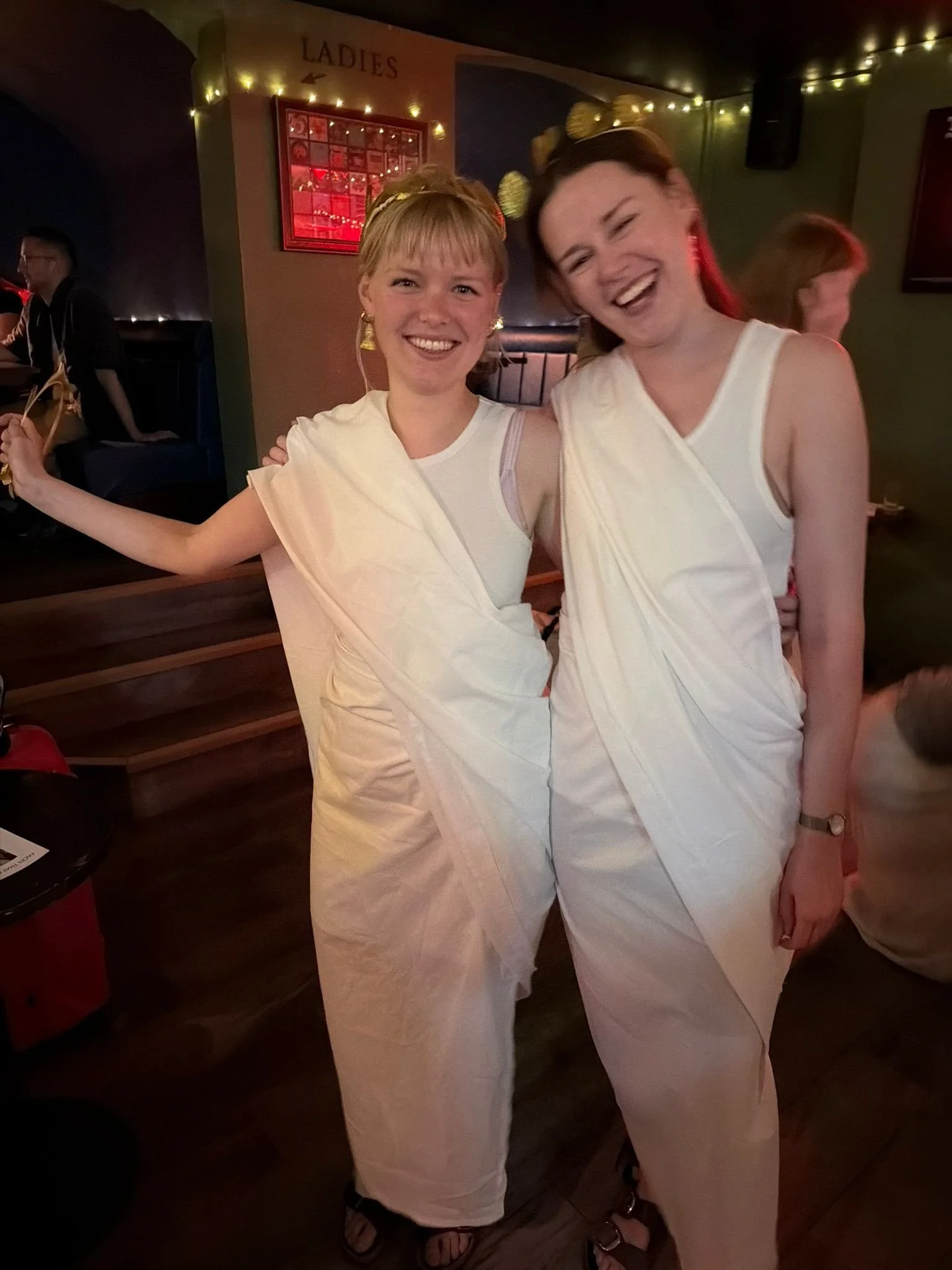 Two women dressed in white toga costumes with gold accessories, smiling at a party or celebration.