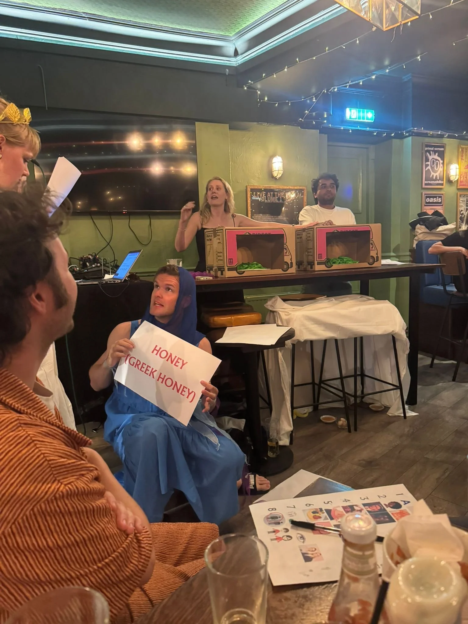 People sitting around a table in a restaurant or bar, some holding signs that read "Honey Greek Honey." There are boxes of vegetables or fruits on the bar, and posters on the walls behind. The people appear to be engaged in conversation or an event.