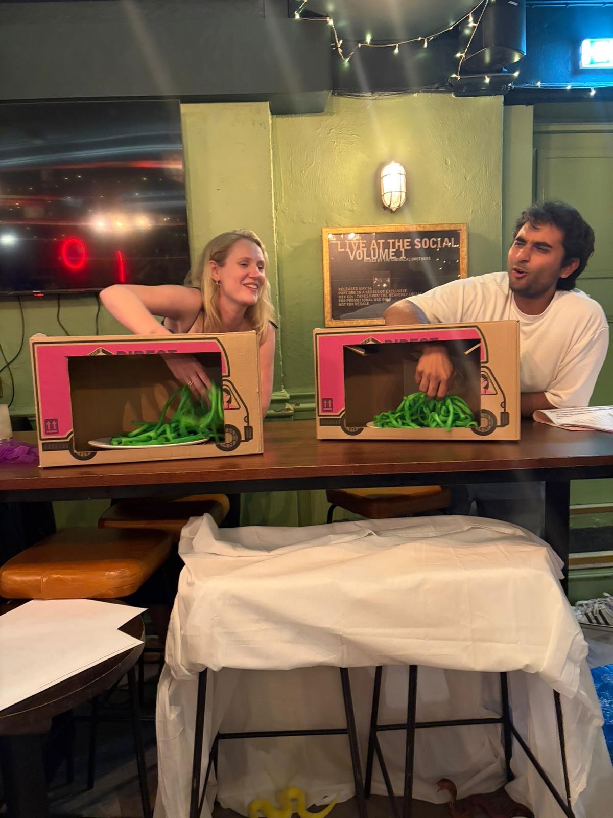 Two people are seated at a bar with cardboard boxes labeled as delivery trucks in front of them. The woman on the left is smiling and reaching into her box, which contains green pasta noodles. The man on the right is also reaching into his box of gre