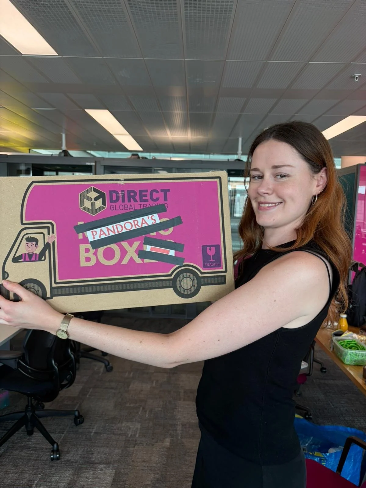 A woman with red hair smiling while holding a cardboard box labeled 'Pandora's Box' with some black tape over part of the label, inside an airport terminal.