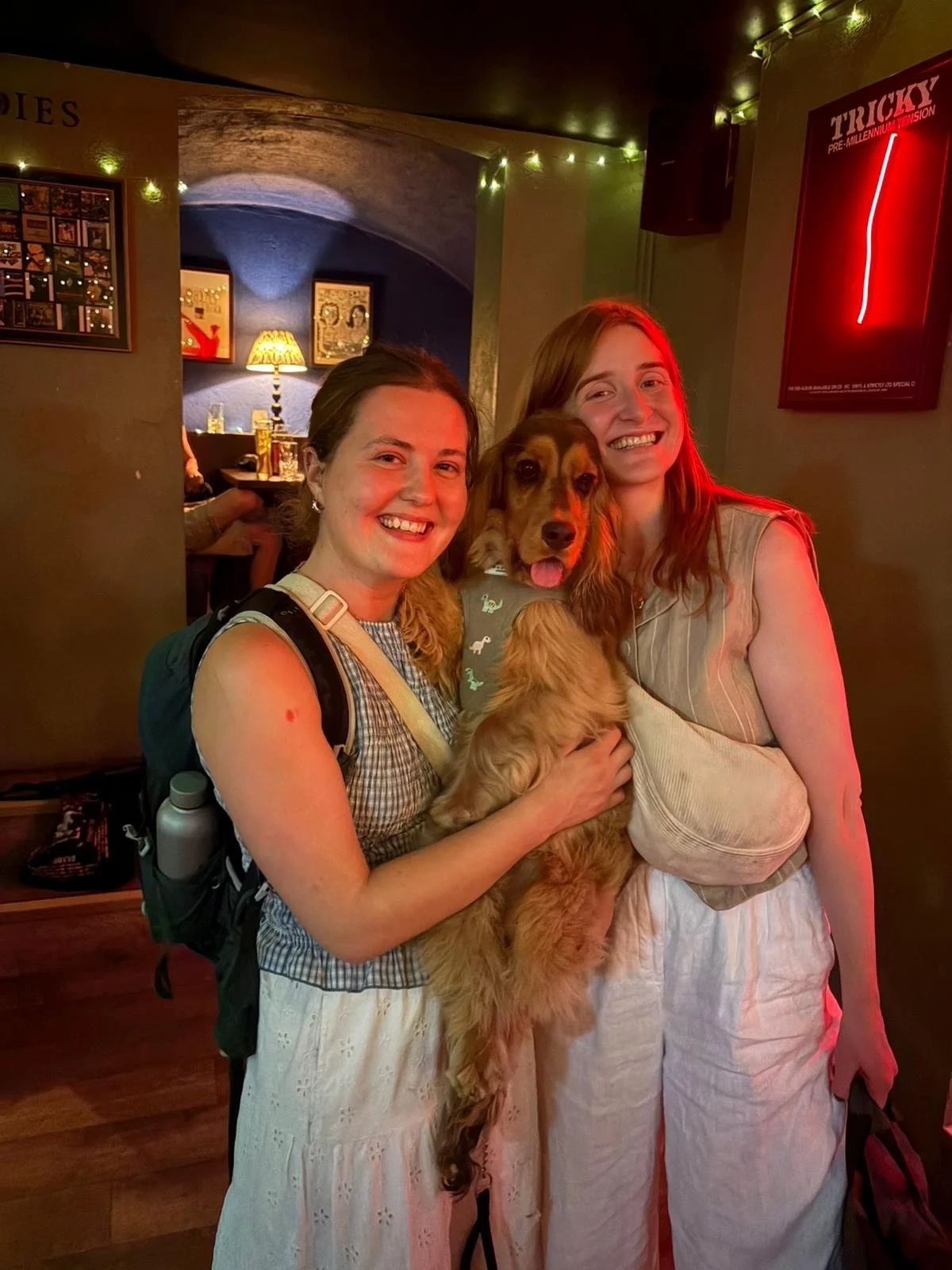 Two women smiling and holding a dog in a dimly lit indoor setting, with warm and colorful lights.