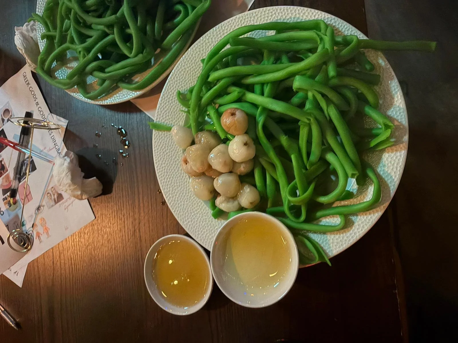 Plate of cooked green beans with pickled garlic and two cups of tea on a wooden table, with some scattered decorations and napkins nearby.