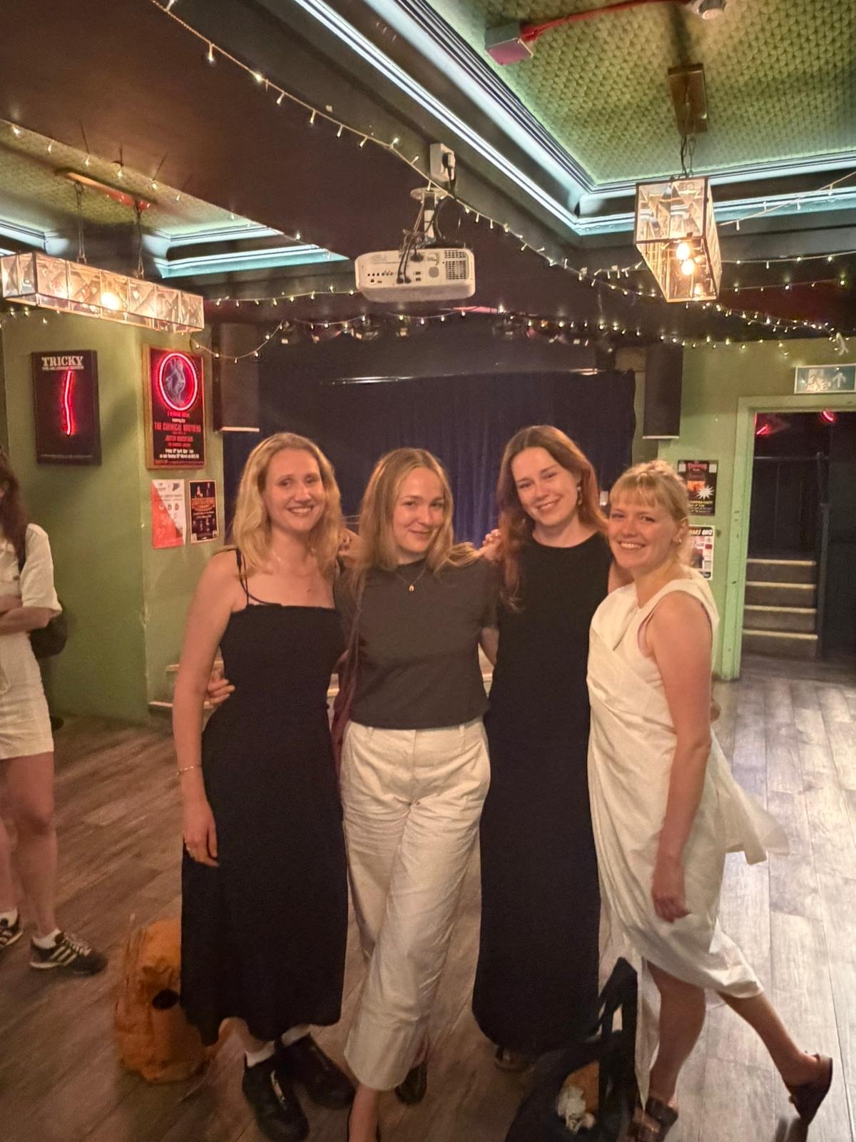 Four women standing together in a dimly lit room, smiling for the photo. The room has string lights, a projector, framed posters on the wall, and a dark stage area in the background, suggesting a social or entertainment venue.