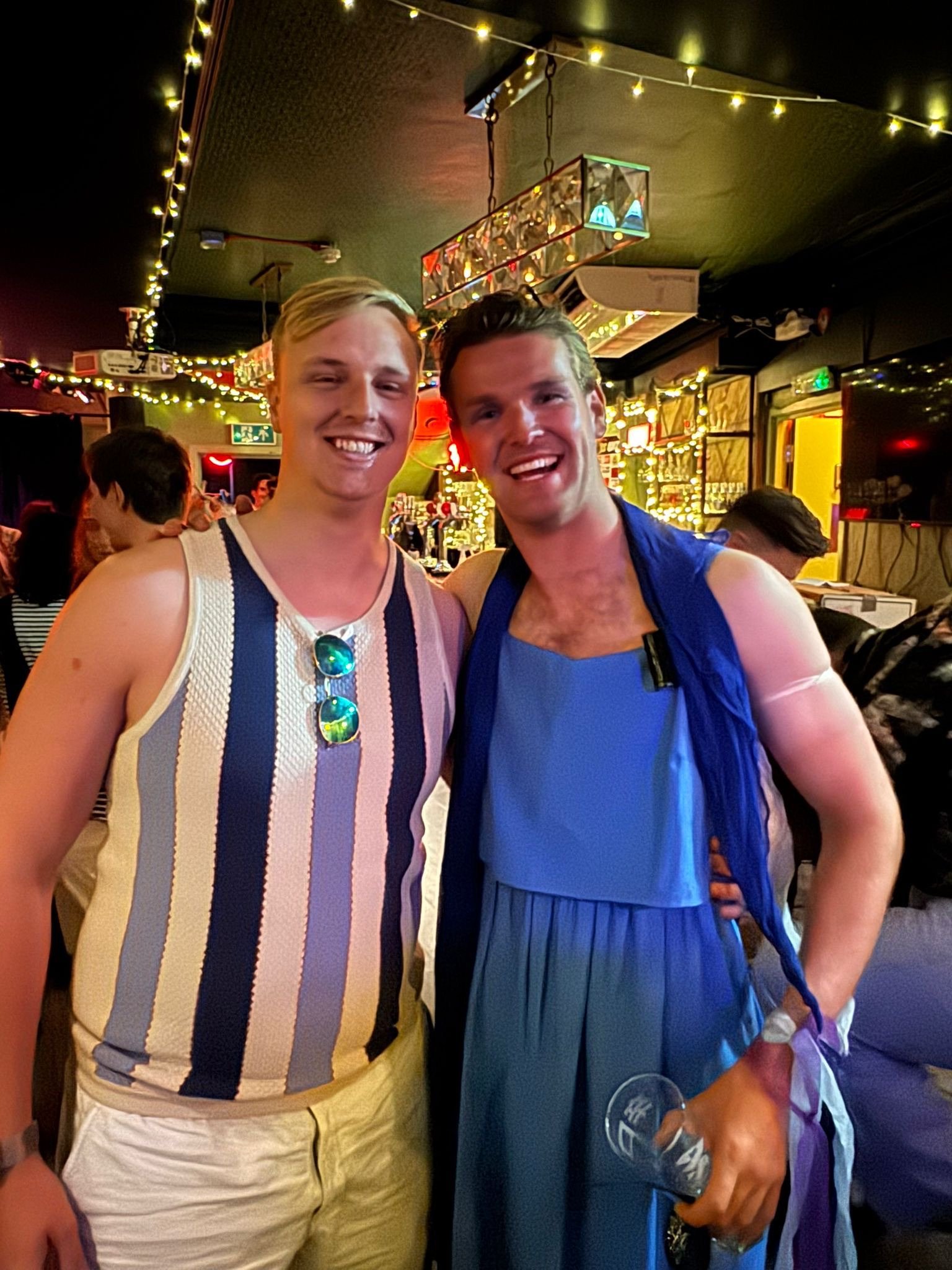 Two young men smiling and posing together in a bar or pub with string lights and various decorations on the walls in the background.