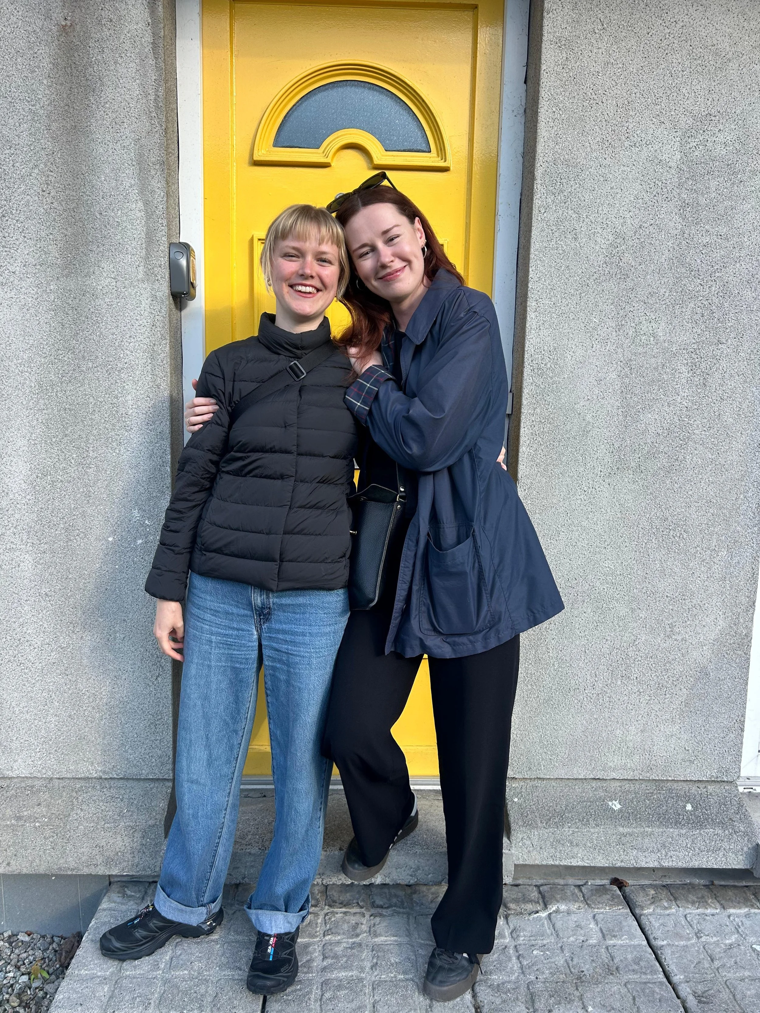 Two women smiling and hugging in front of a yellow door. One wears a black puffer jacket and jeans, the other a dark blue jacket and black pants.