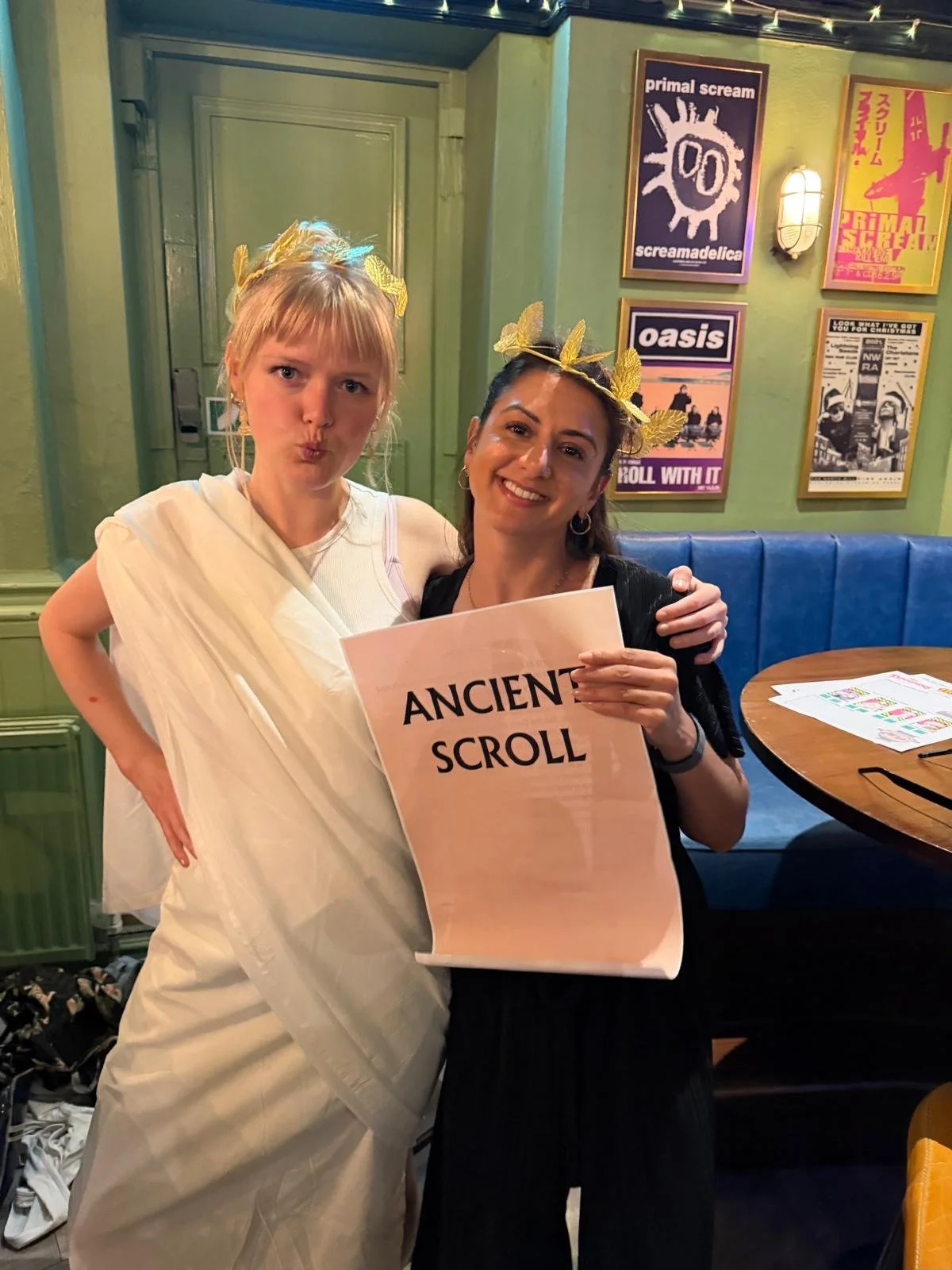 Two women, one dressed as a Greek goddess and the other holding a sign saying "Ancient Scroll," standing inside a colorful restaurant with musical posters on the wall, a round table, and string lights overhead.