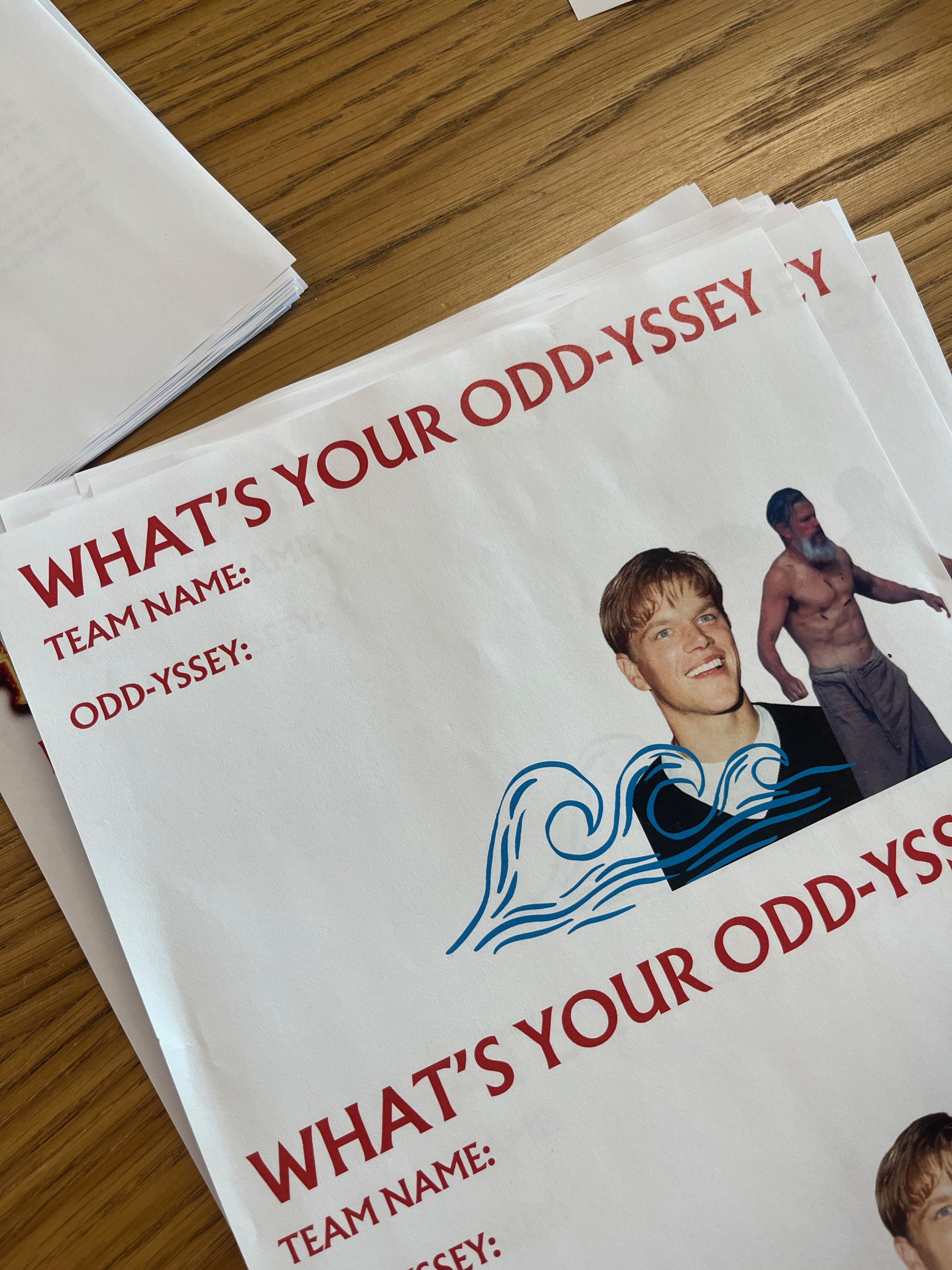 A printed info sheet with the title 'WHAT'S YOUR ODD-YSSY?', partially visible, featuring images of people, including a young man smiling and an older man with gray hair and a beard, shirtless, with waves and water graphic design on the sheet, placed on a wooden surface.