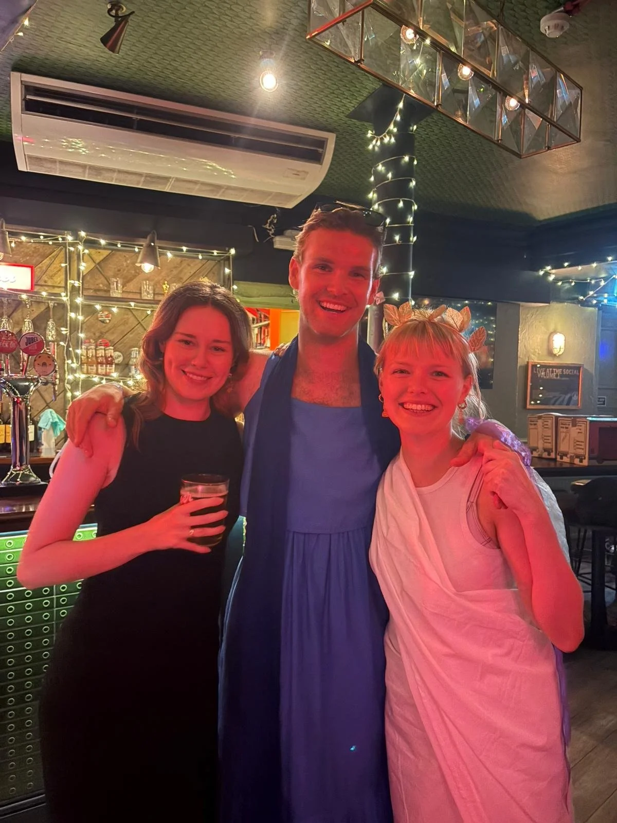 Three friends smiling and posing together in a dimly lit bar or pub, with fairy lights and bar decor in the background.