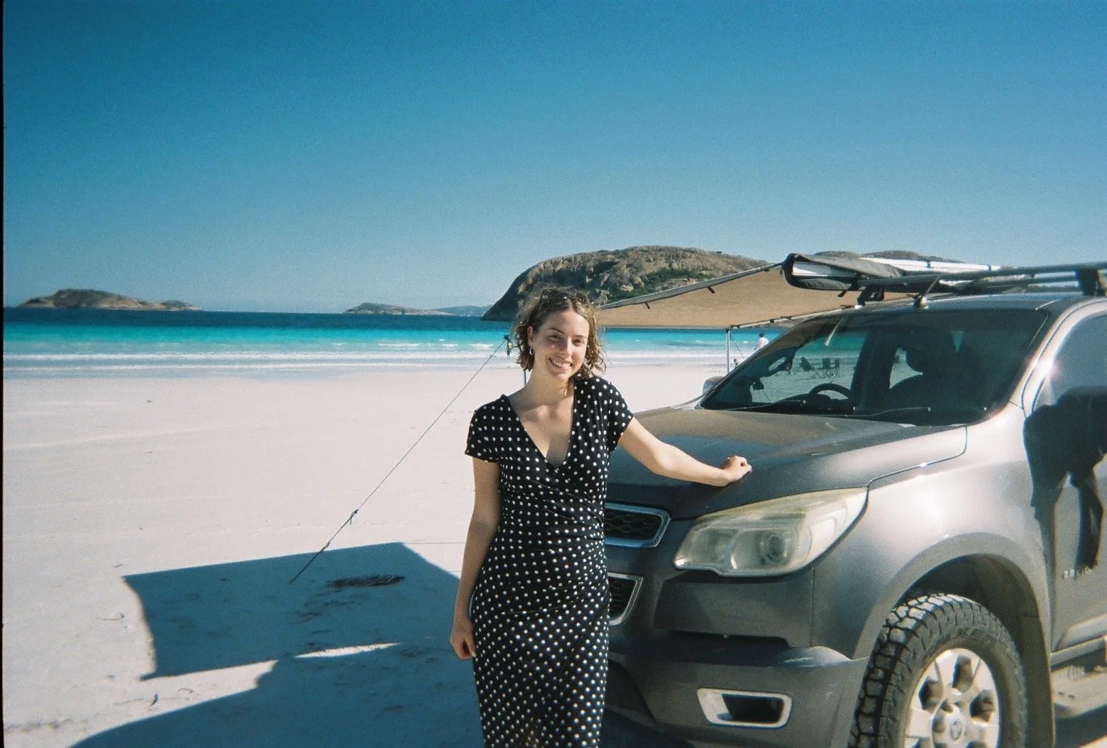 A woman in a black and white polka dot dress standing next to a dark gray SUV on a sandy beach with a large umbrella and small islands in the background.
