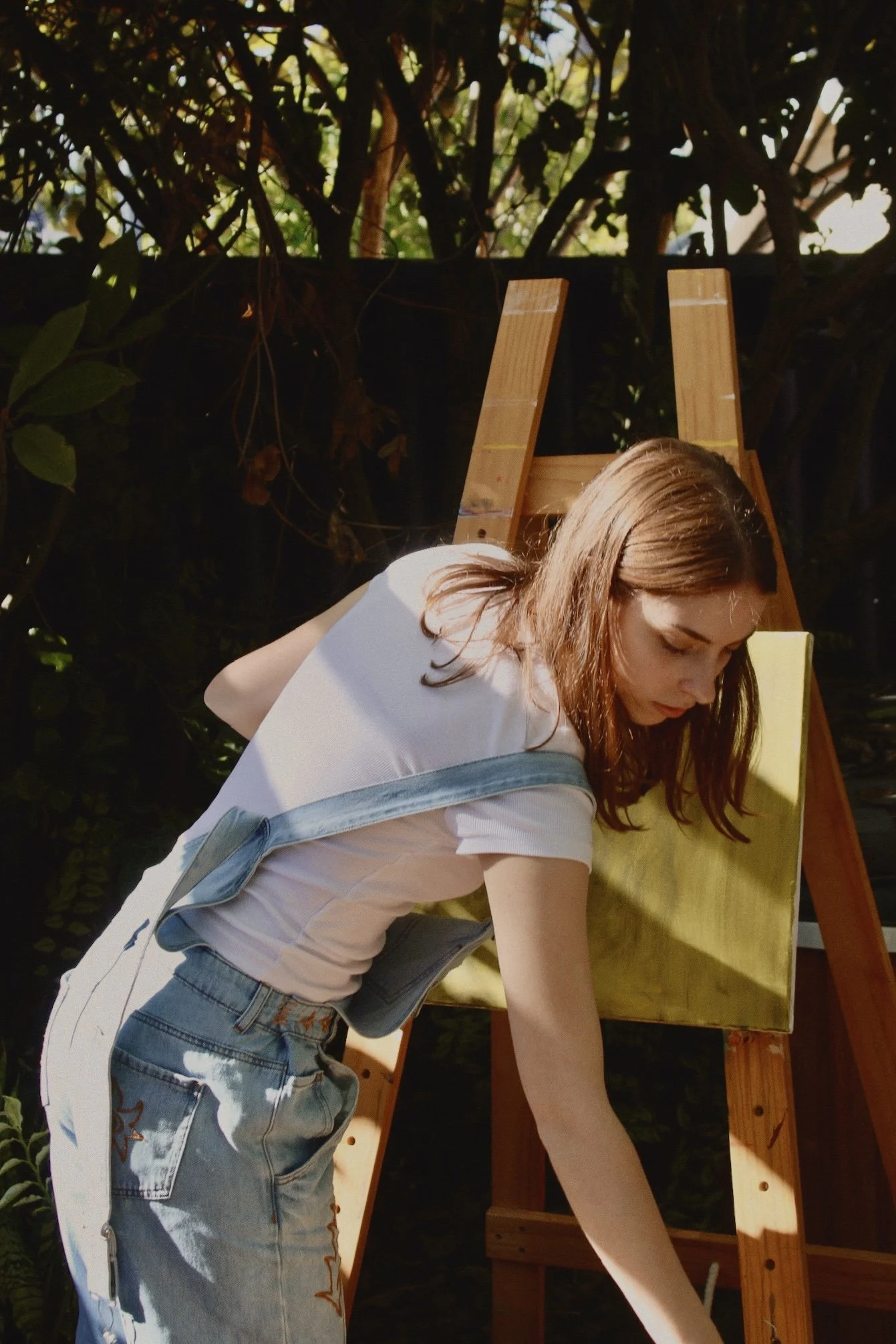 A young woman in a white t-shirt and overalls bending down to pick up a paintbrush. Behind her is a wooden easel with a blank green canvas and foliage, indicating a garden or backyard setting.