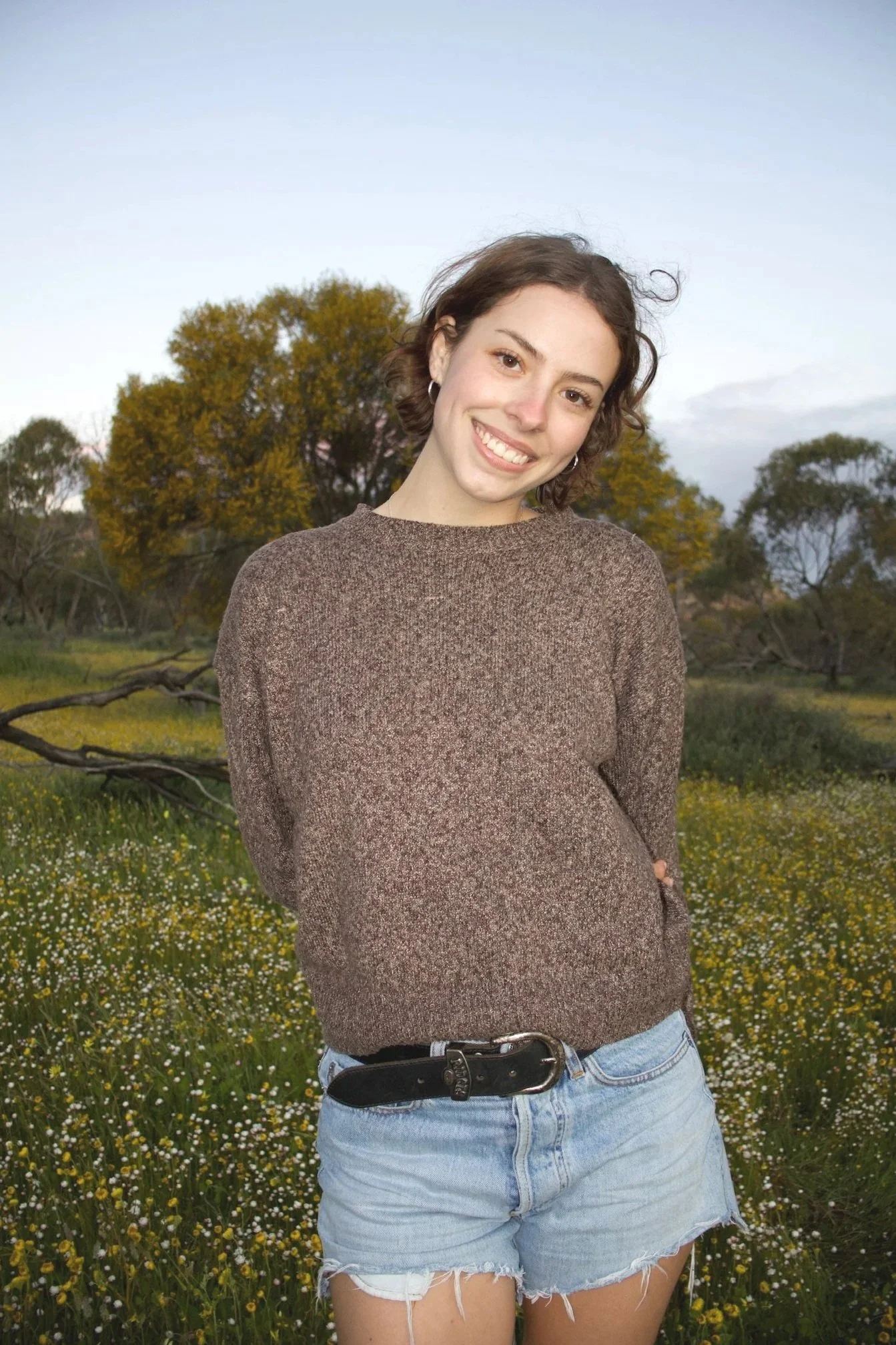 A smiling young woman with brown hair in a chin length bob, wearing a brown sweater and light blue denim shorts, stands in a field of small white and yellow flowers with trees in the background under a partly cloudy sky.