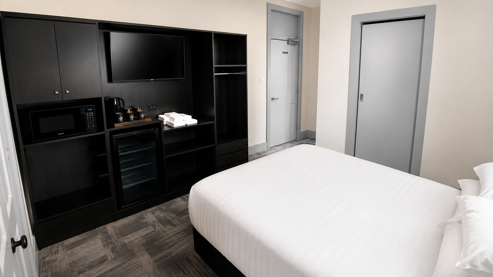 Hotel room with a bed, black entertainment center with TV, microwave, mini fridge, coffee kettle, and towels, neutral walls, gray doors.