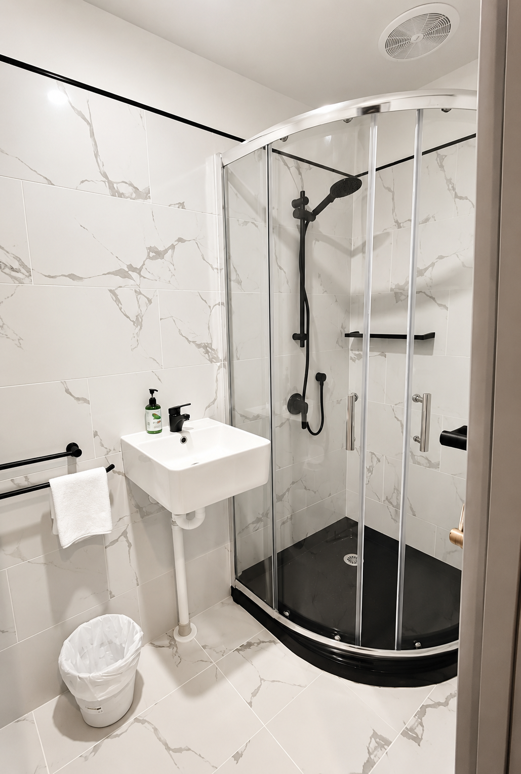 Small modern bathroom with marble tiles, corner shower with glass doors, black shower fixtures, small sink, black soap dispenser, towel on rack, trash can, and ceiling vent.