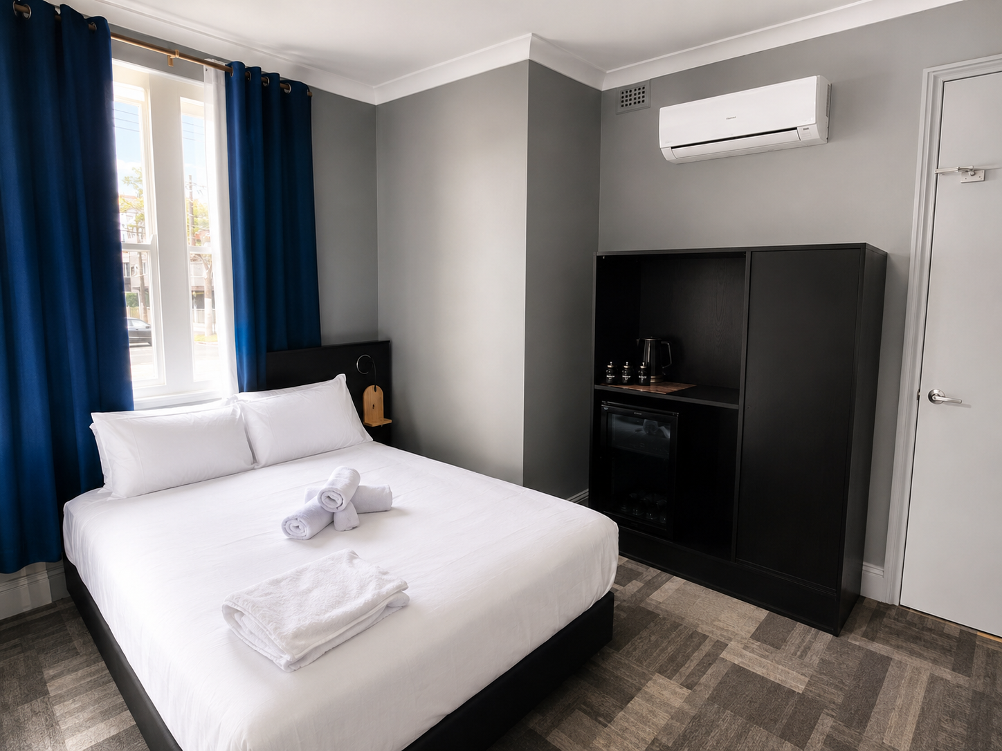 A hotel room with a bed, blue curtains, a window, a black cabinet with a mini refrigerator and a coffee station, an air conditioning unit, and a closed white door.