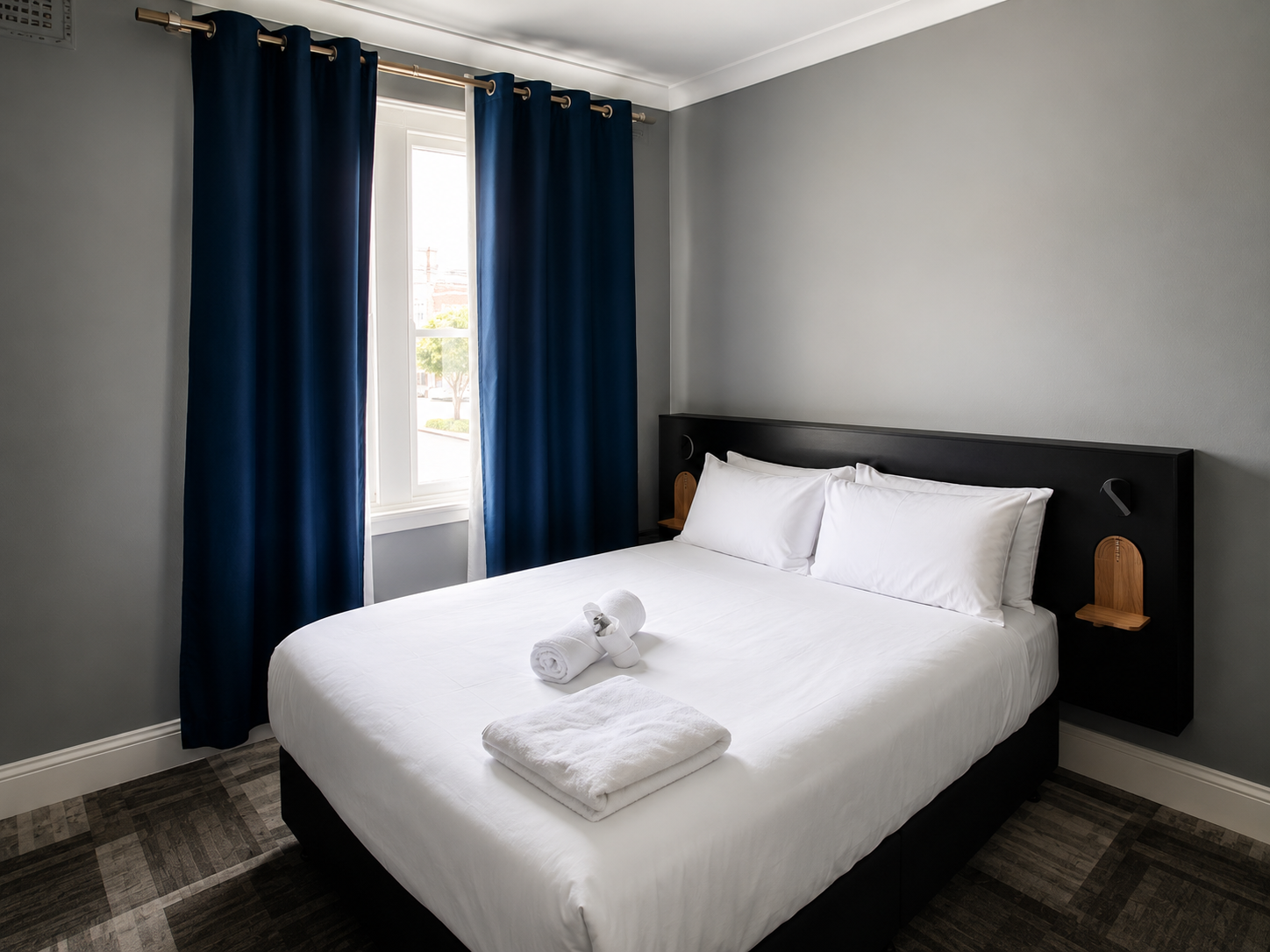 A neatly made bed with white linens and two pillows, in a room with gray walls, blue curtains, and a window letting in natural light.