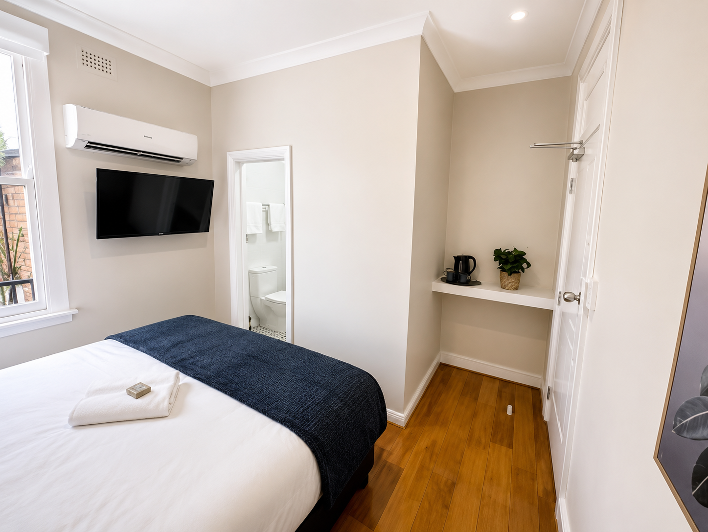 A bedroom featuring a bed with a white sheet and a dark blue blanket, a mounted flat-screen TV, a window, a small wall-mounted shelf with a potted plant and a coffee maker, and a partial view of a bathroom with a toilet and towels.