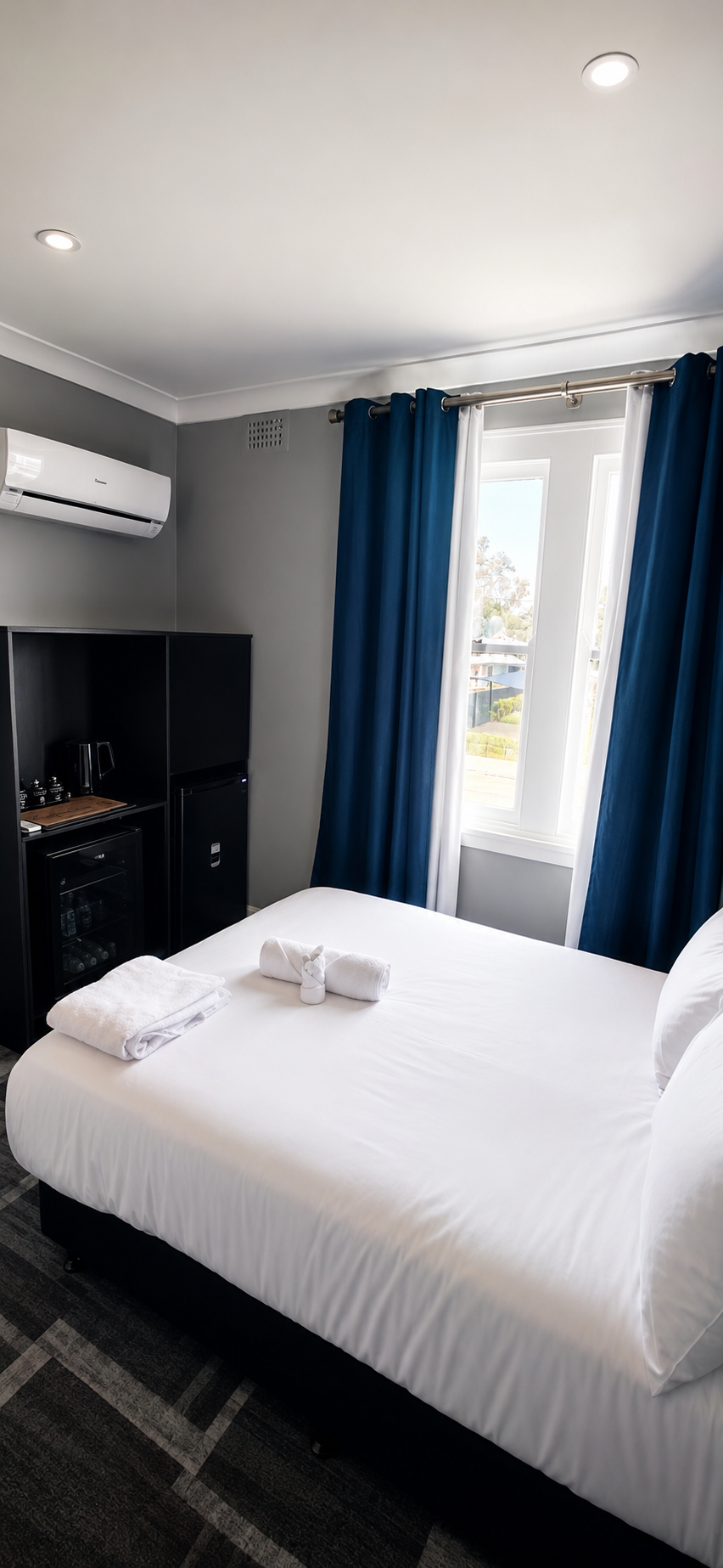 Hotel room with white bed, blue and white curtains, window with a view outside, black cabinet with mini fridge, towels and toiletries on the bed, and ceiling lights.