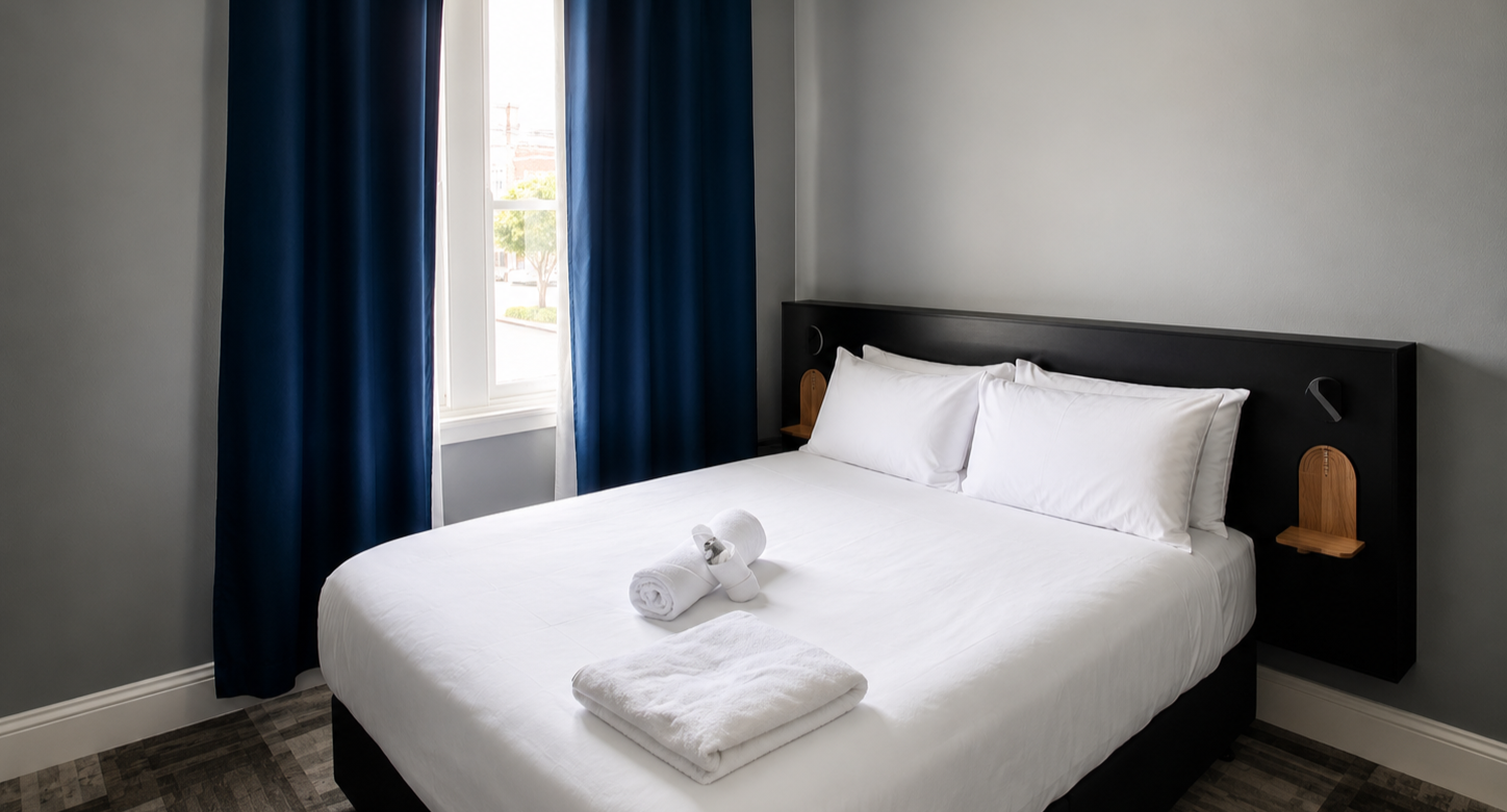 A neatly made hotel room bed with white linens, pillows, and rolled towels on top, situated near a window with blue curtains.