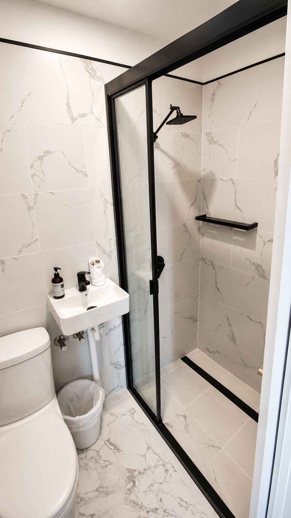 Small modern bathroom with a toilet, a tiny sink, and a walk-in shower with black frame and marble tiles.