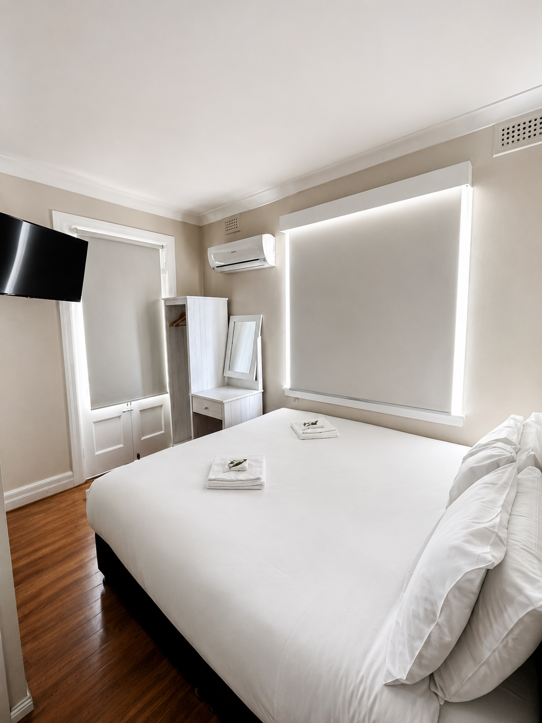 A minimalistic hotel room with a large bed, white bedding, two neatly folded towels with small decorative sprigs, two pillows, a small white wardrobe with a mirror, a wall-mounted TV, a window with blinds, and a wooden floor.