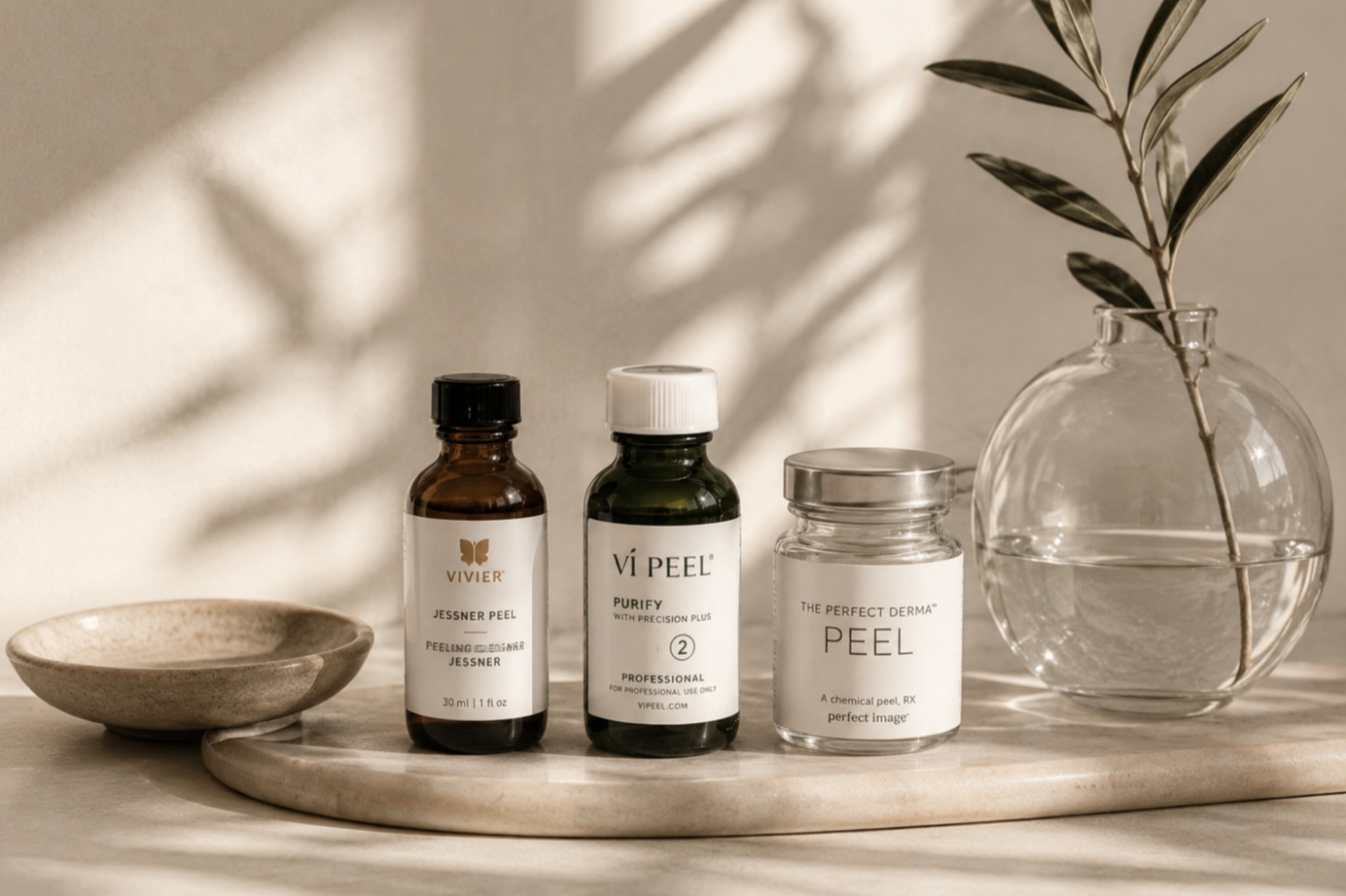 Skincare products, including peel bottles and jars, on a marble surface next to a glass vase with a branch and leaves, with soft shadows in the background.