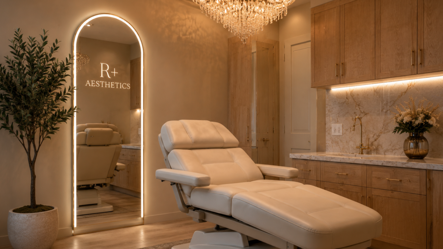 A comfortable medical or aesthetic treatment room featuring a white reclining chair, a tall decorative mirror with branding that reads 'R+ Aesthetics,' a potted plant, warm wooden cabinetry, and a chandelier.