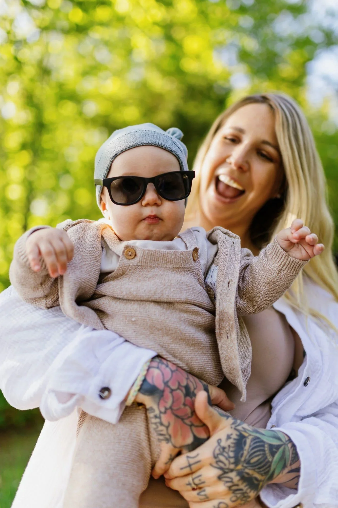 Baby mit Sonnenbrille und Mütze auf dem Arm der lachenden Mutter im Sommer.