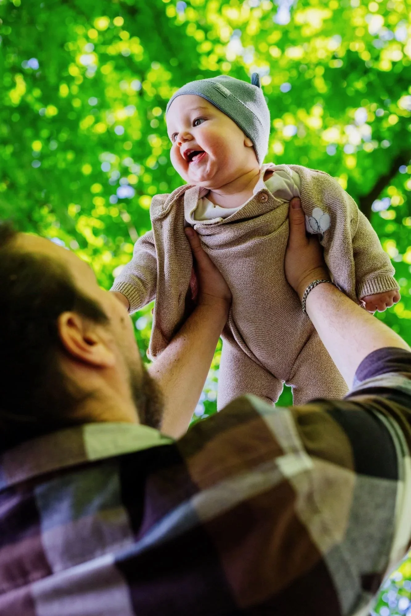 Ein Baby wird in einem Park von einer Person hochgehoben, umgeben von grünen Blättern im Hintergrund.