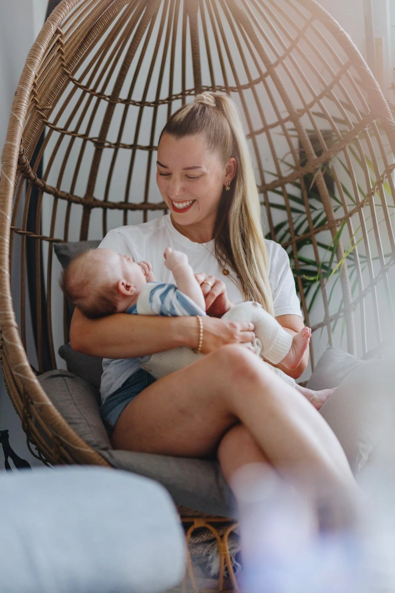 Lachende Mutter hält ihr Baby im Arm in einem Hängesessel bei Tageslicht, natürlich und unposed.