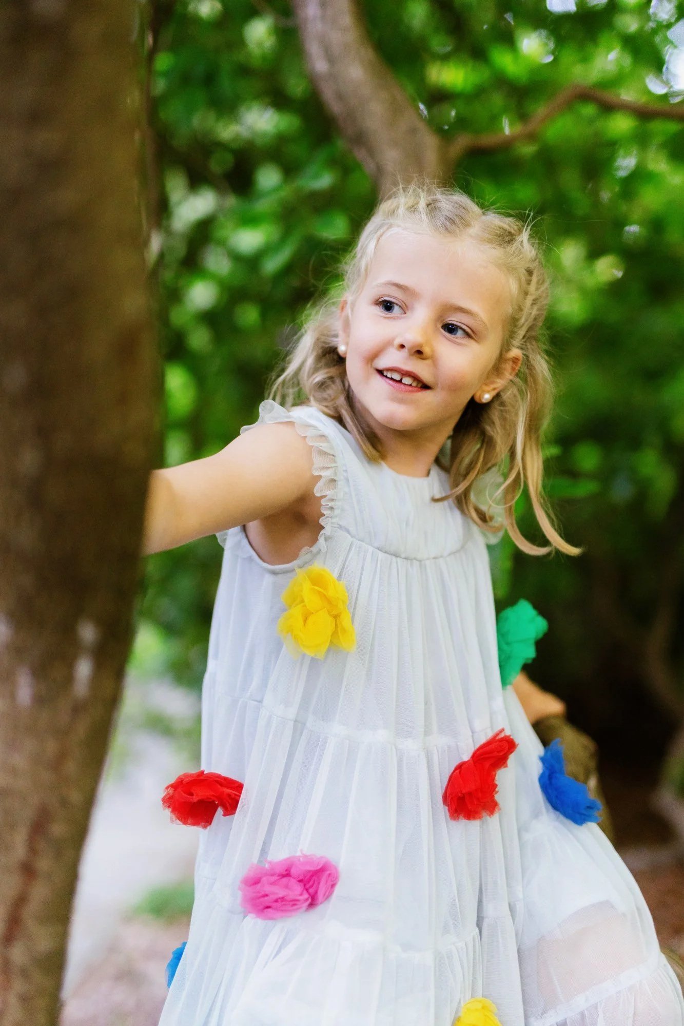 Lächelndes Mädchen im weißen Kleid mit bunten Blüten lehnt an einem Baum.