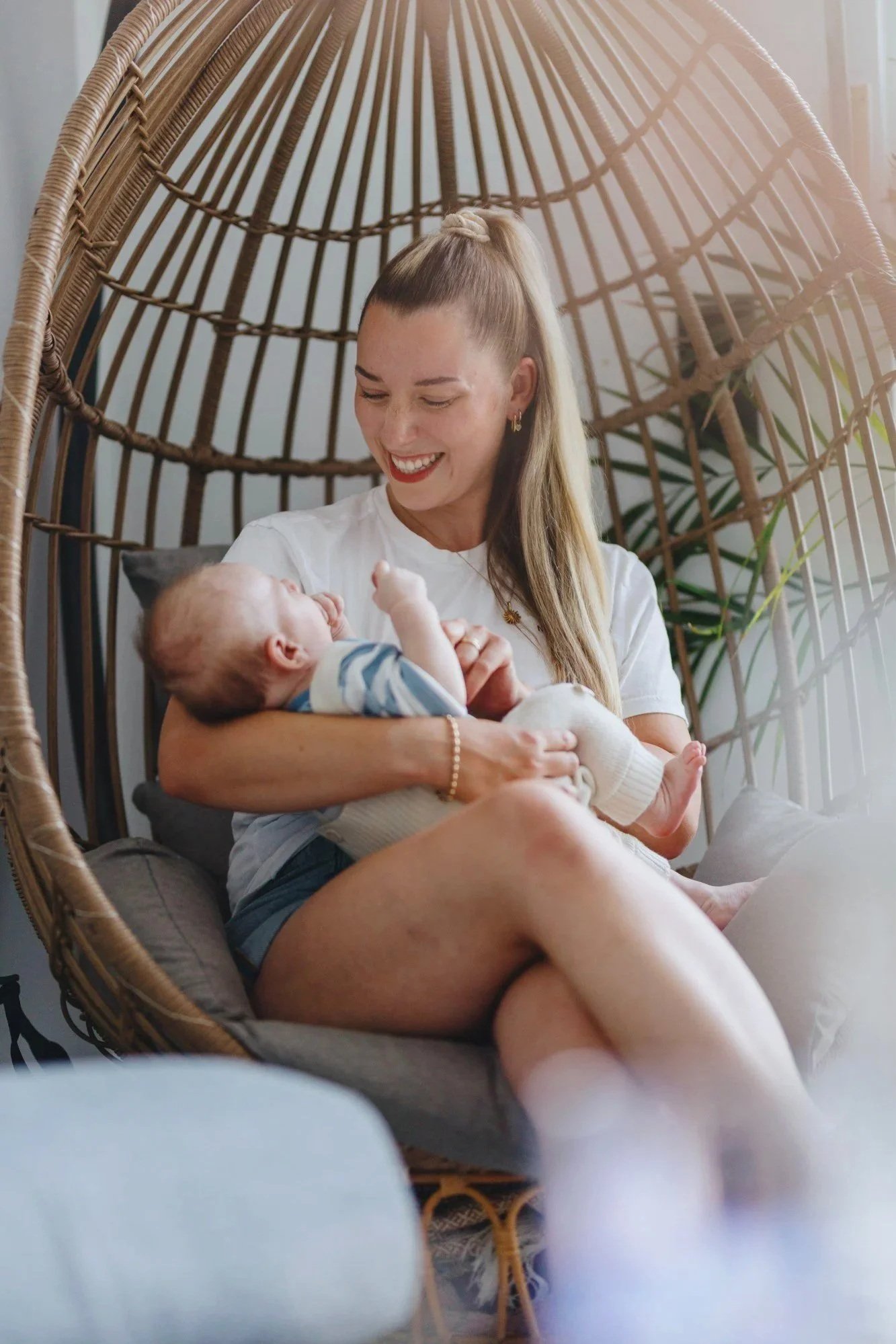 Lachende Mutter hält ihr Baby im Arm in einem Hängesessel bei Tageslicht, natürlich und unposed.