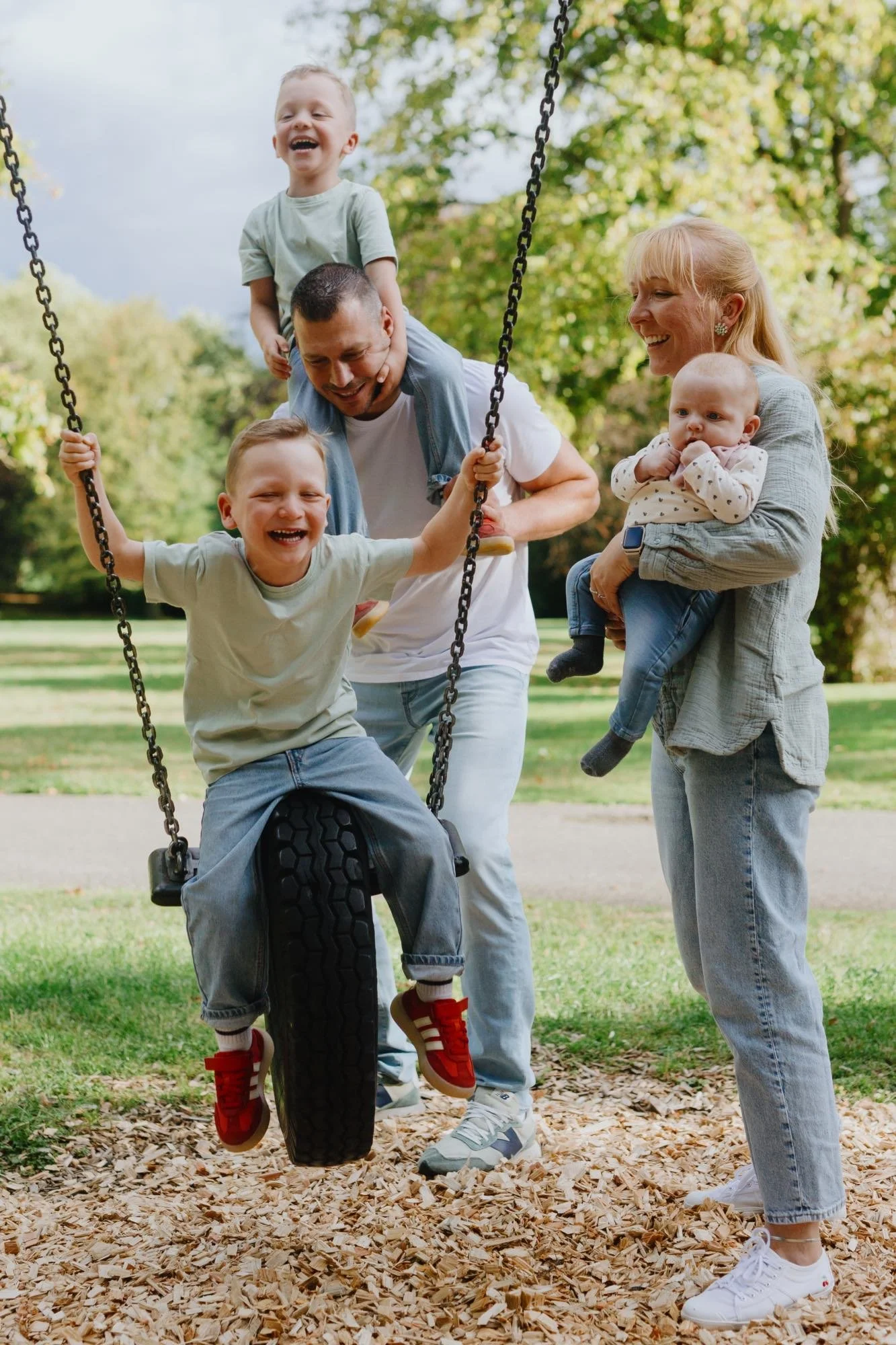 Fünfköpfige Familie lacht gemeinsam auf dem Spielplatz; ein Junge sitzt auf der Reifenschaukel.