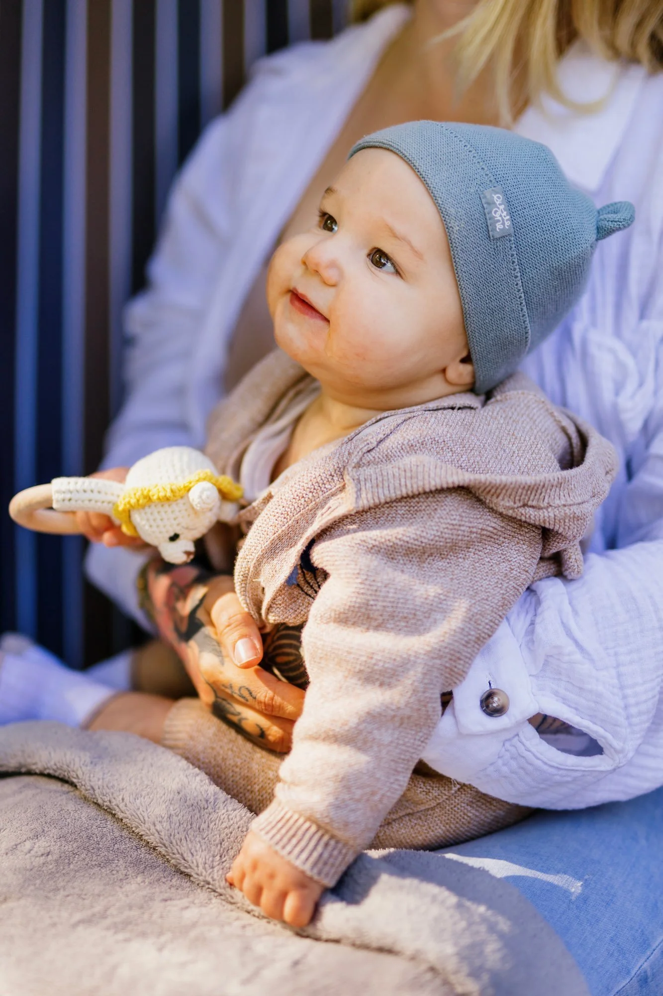 Baby mit Mütze sitzt auf dem Schoß und hält eine gehäkelte Rassel im warmen Licht.