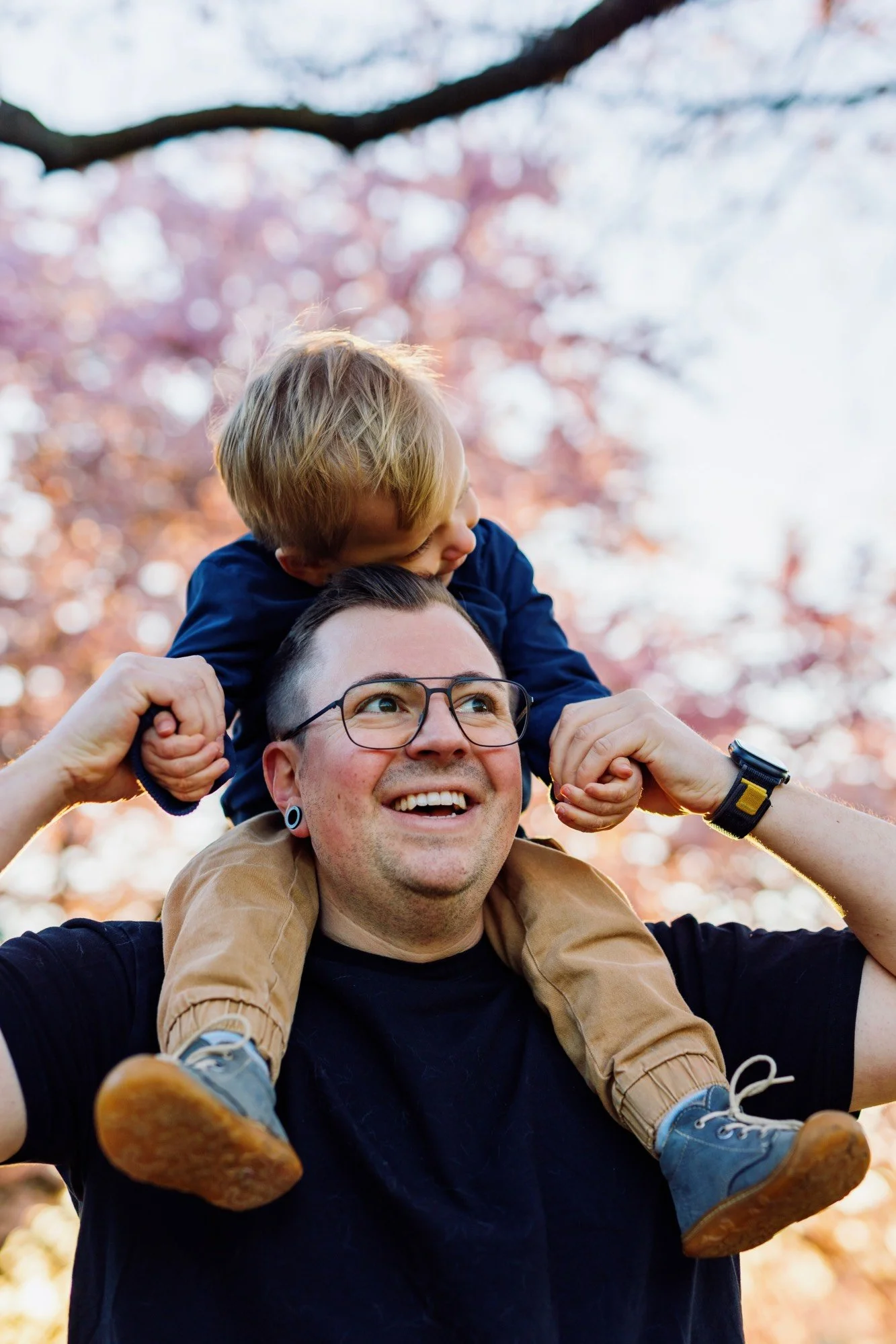 Lachender Vater trägt sein Kind auf den Schultern unter blühenden Kirschbäumen, Familienfotografie im Frühling.