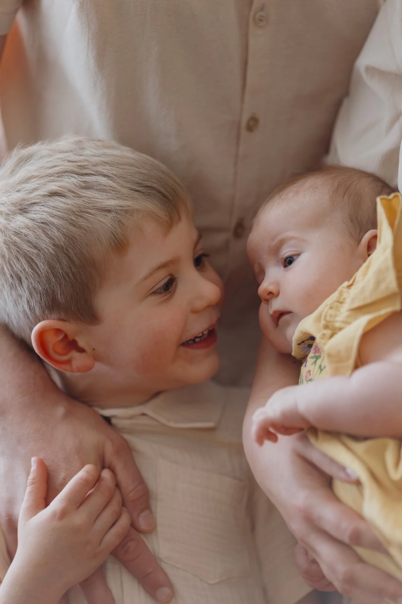 Zwei Kinder, ein älterer Junge und ein neugeborenes Baby, schauen sich liebevoll an und werden von einer erwachsenen Hand gehalten, die sie nahe beieinander hält.