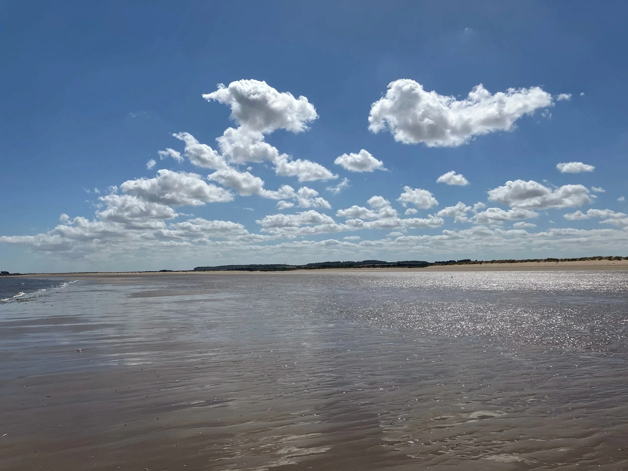 Wet sand and shallow water on Titchwell beach beneath a blue sky with scattered clouds.