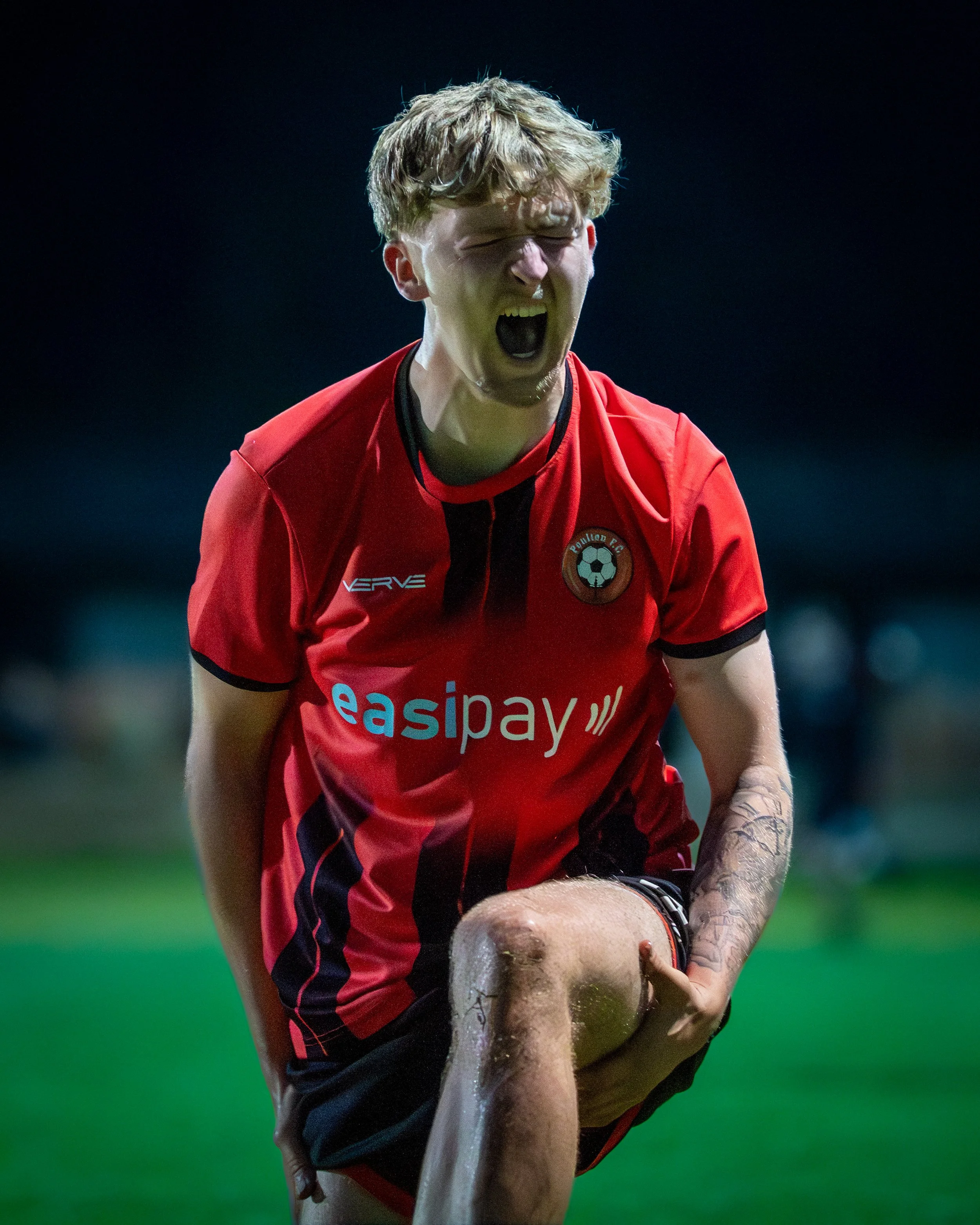 A soccer player in a red uniform with a logo reading 'Raispay' and a badge with a soccer ball, yells in excitement or frustration during a game, with his eyes closed, steam or sweat visible on his face, and tattoos on his left arm.
