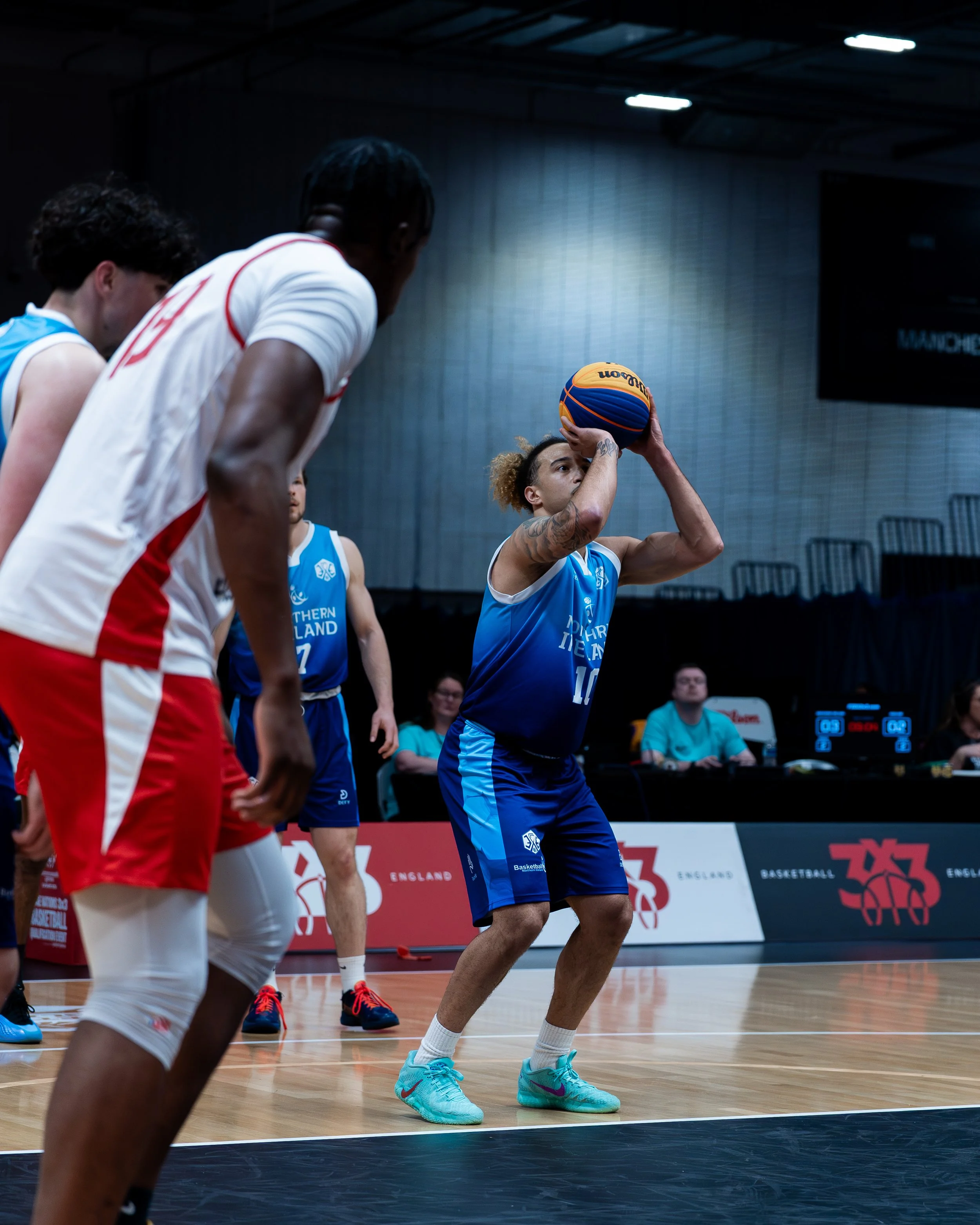 Basketball player preparing to shoot in a game, wearing a blue uniform, in an indoor gymnasium with spectators and scoreboard in the background.