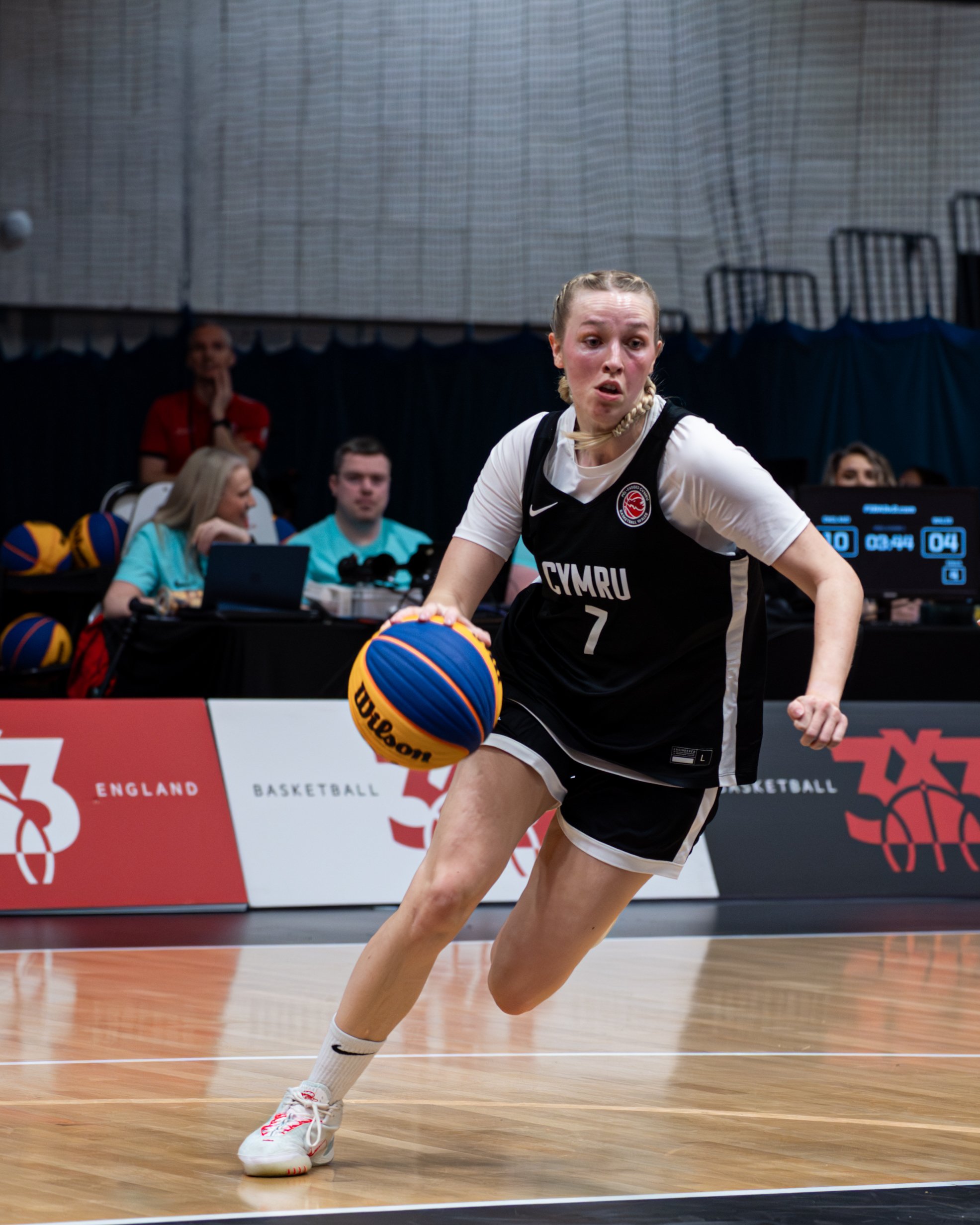 A female volleyball player in a black and white uniform with 'Cymru' written on it, is in a gymnasium falling forward to hit a blue and yellow volleyball. There are other players and officials in the background, with a red and white banner displaying the words 'England Basketball'.